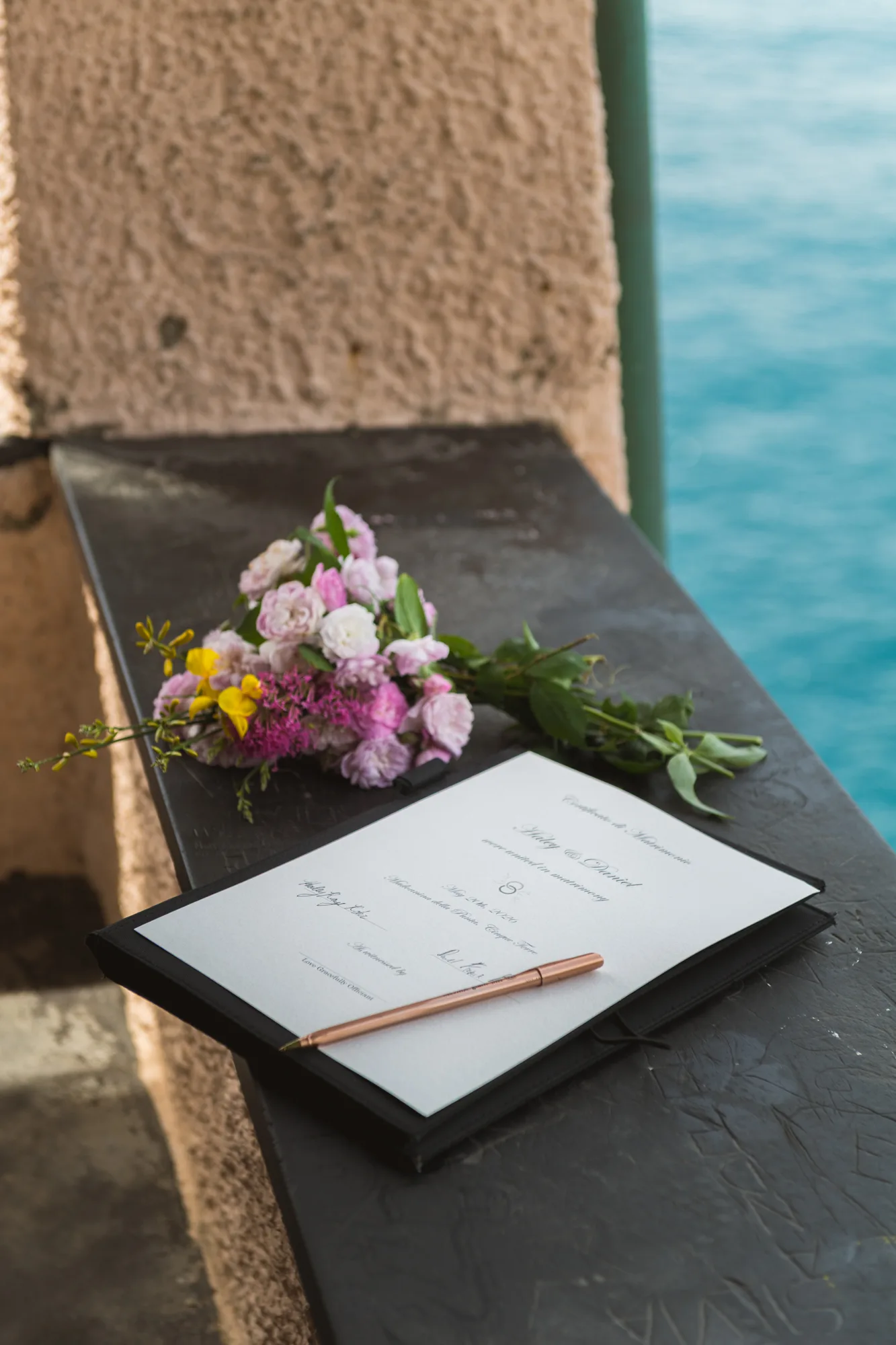The photograph shows a black book or notebook with a pen and a bouquet of flowers placed on top.