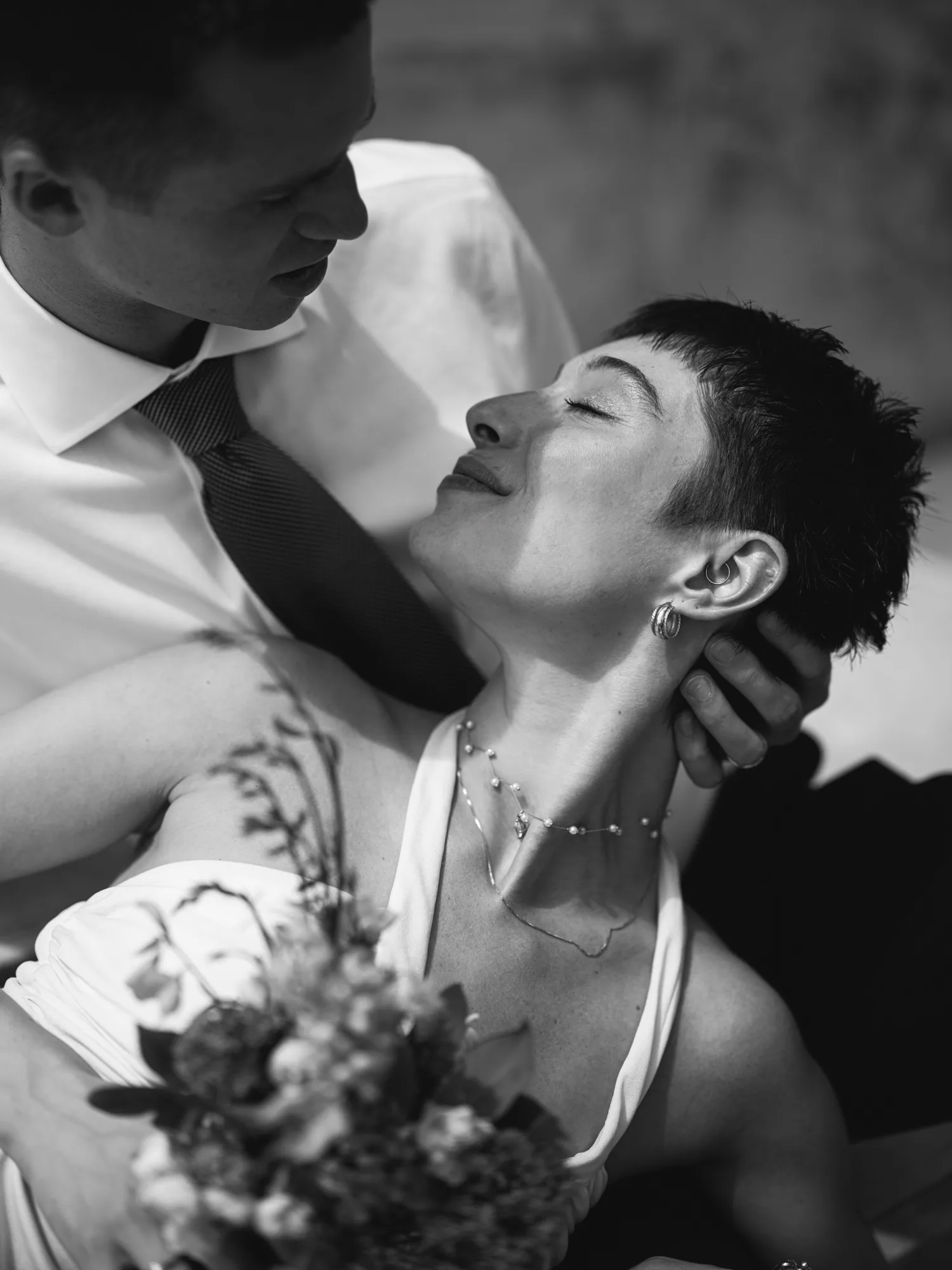 The photograph is in black and white, featuring a close-up of a bride with short hair, wearing a white sleeveless top and a delicate necklace.