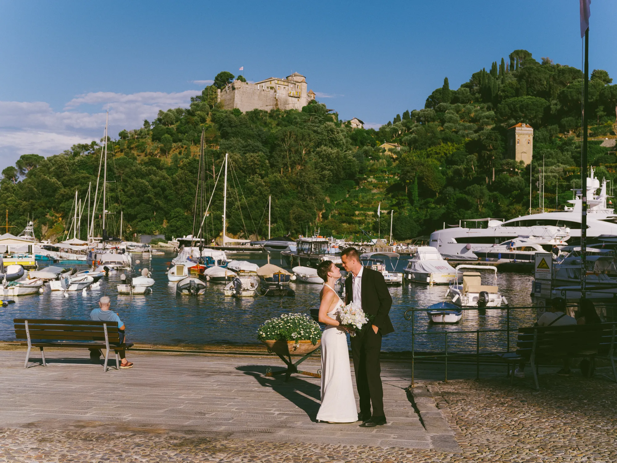 A bride and groom stand on a paved walkway near a marina filled with boats and yachts.