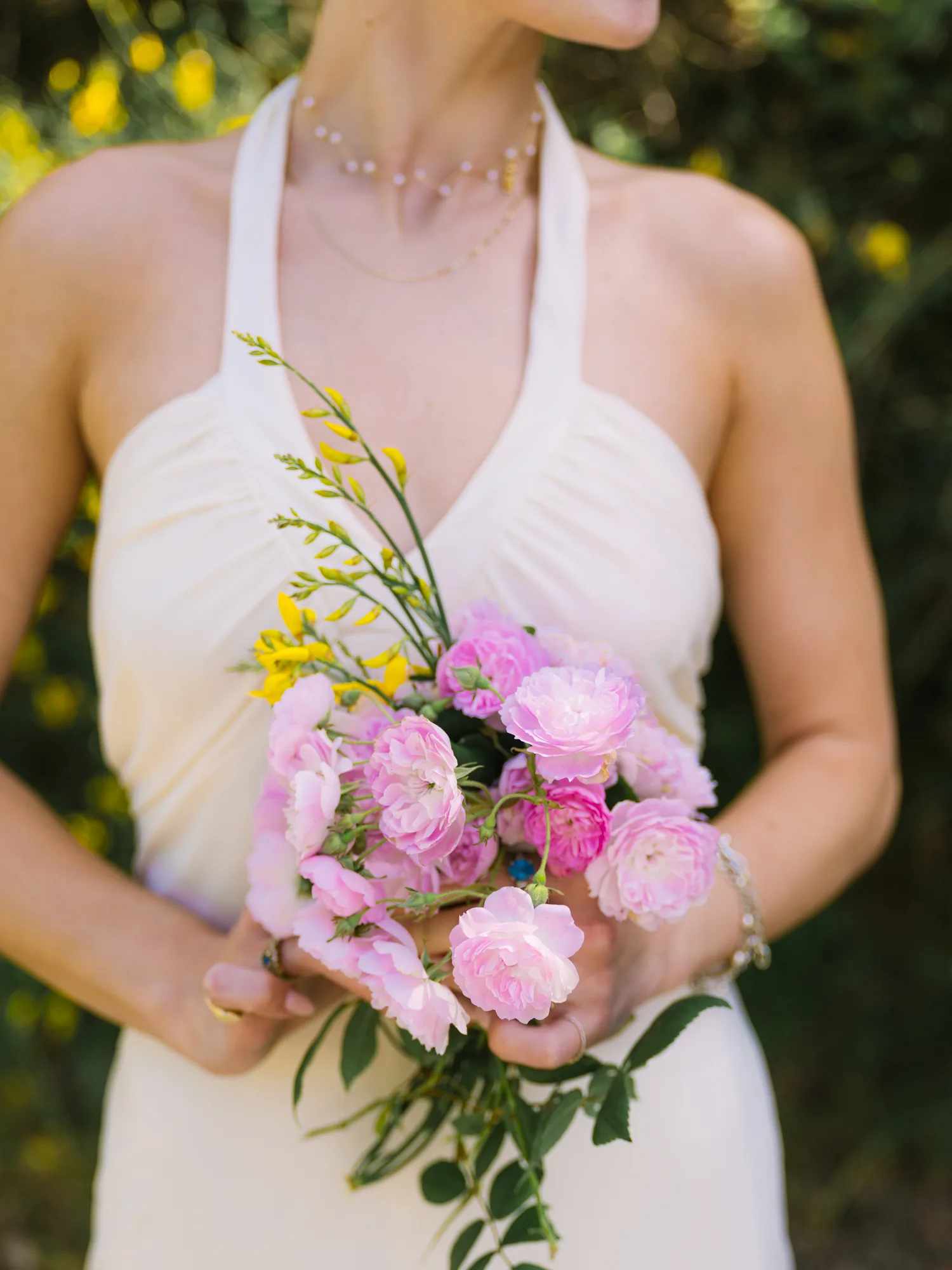 A bride is holding a bouquet of flowers, including pink roses and yellow flowers, in their hands.