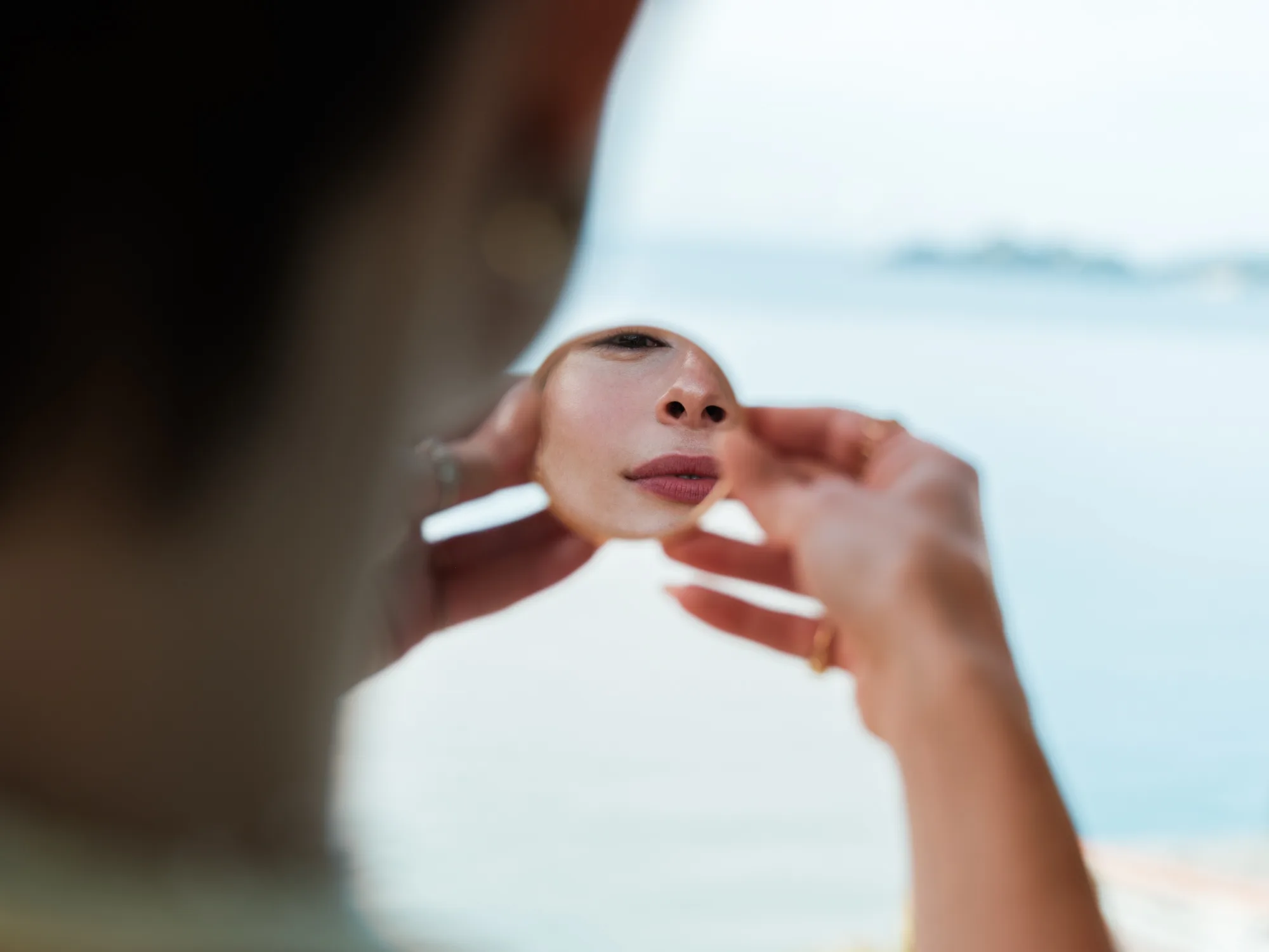 A bride is holding a small round mirror in front of their face, reflecting their nose and part of their cheek.
