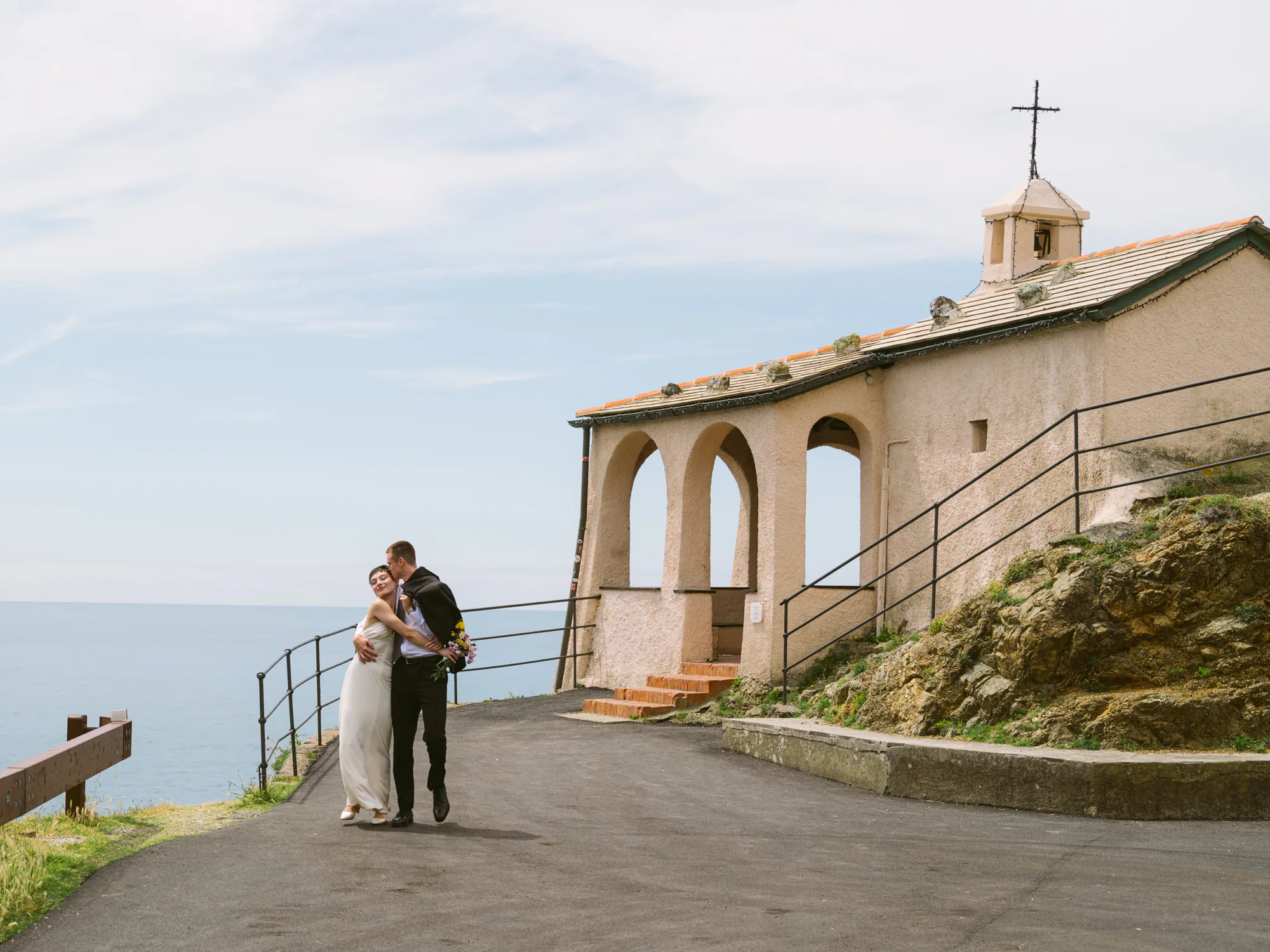 A bride and person stand embracing on a paved path, with the groom in a white dress and the person in a black suit.