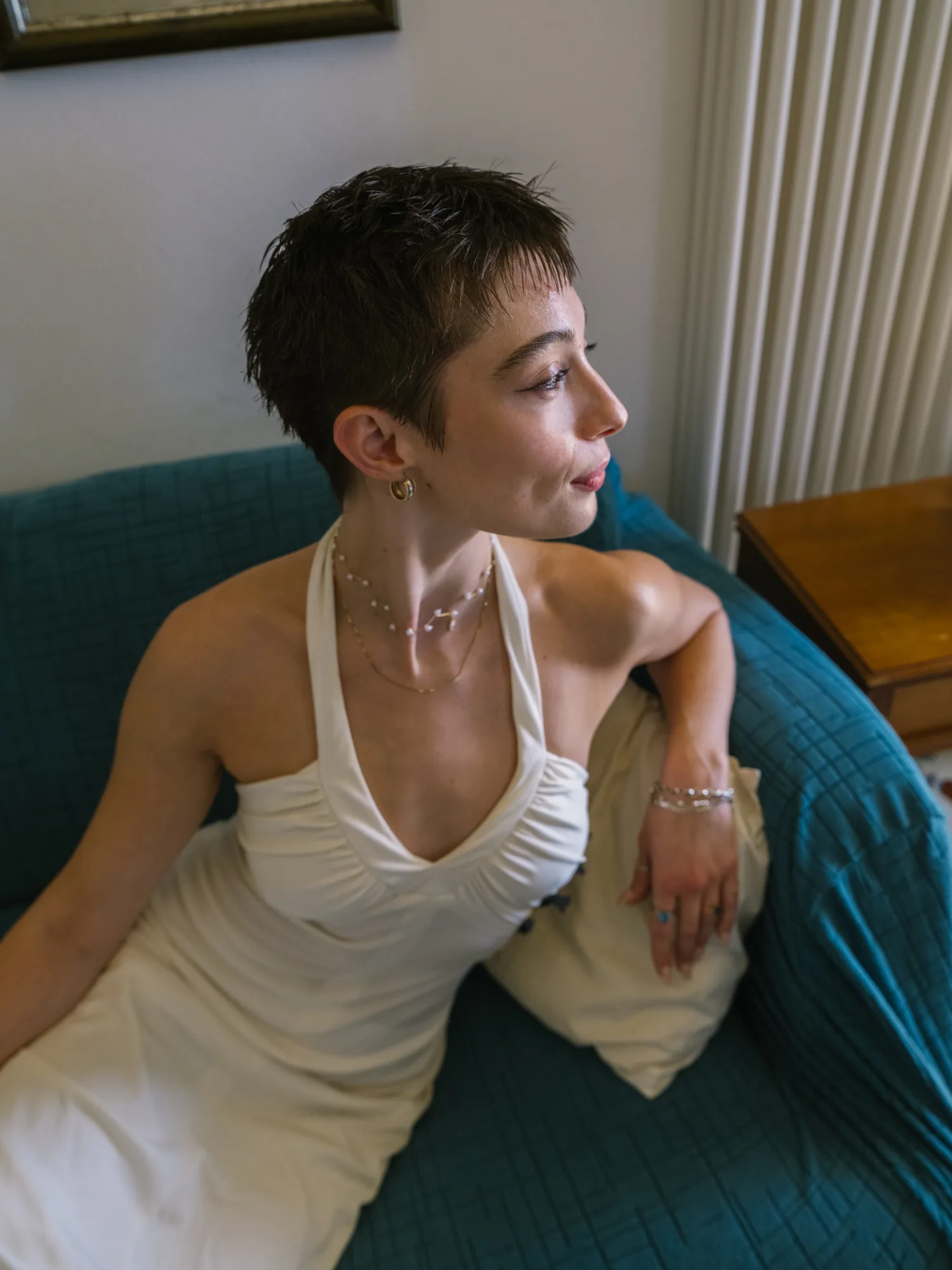 A bride is sitting on a teal couch, wearing a white dress with a one-shoulder design and a delicate necklace.