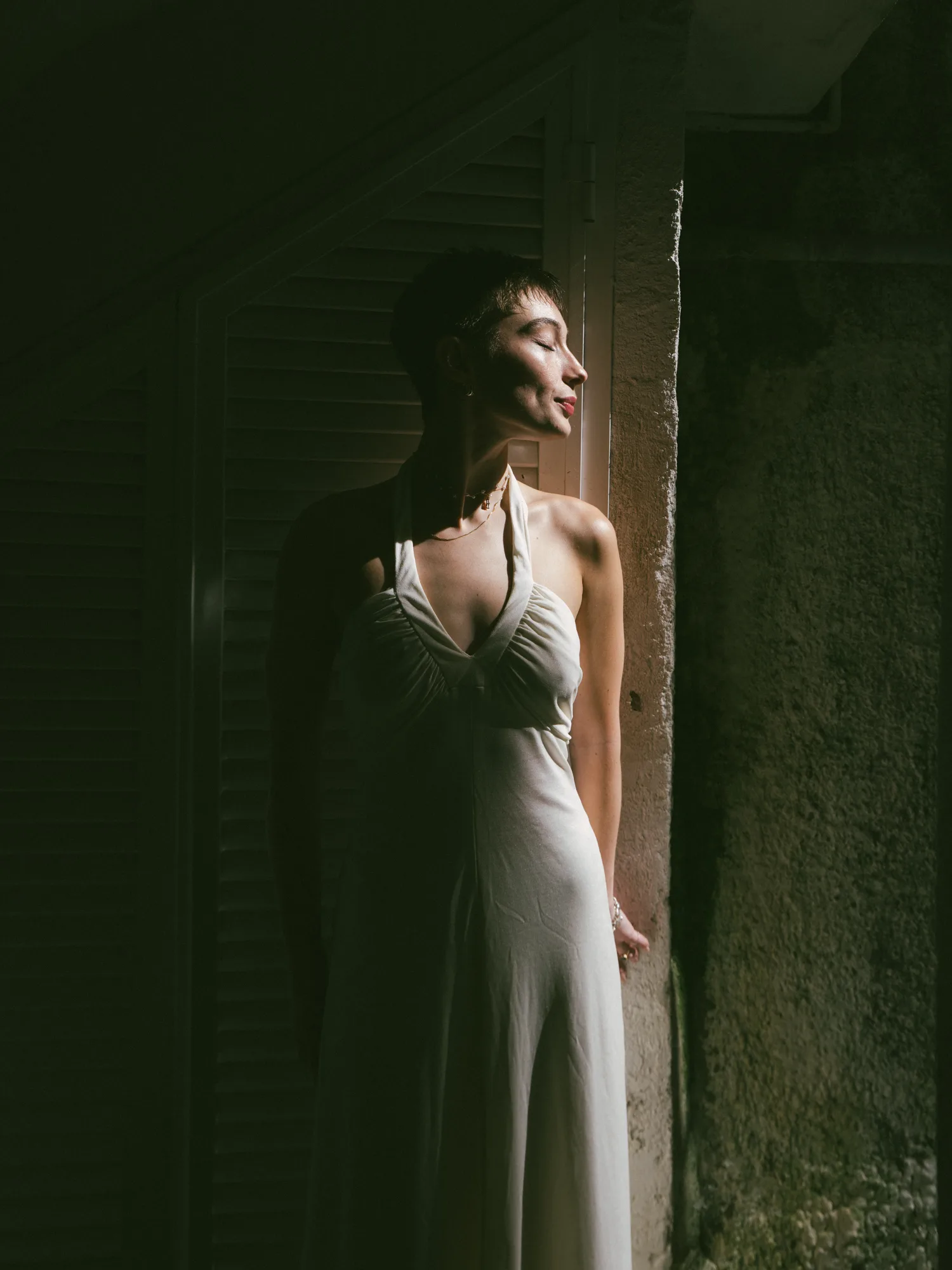 A bride stands in a dimly lit room, wearing a sleeveless, form-fitting dress with a deep neckline and a high slit on one side.