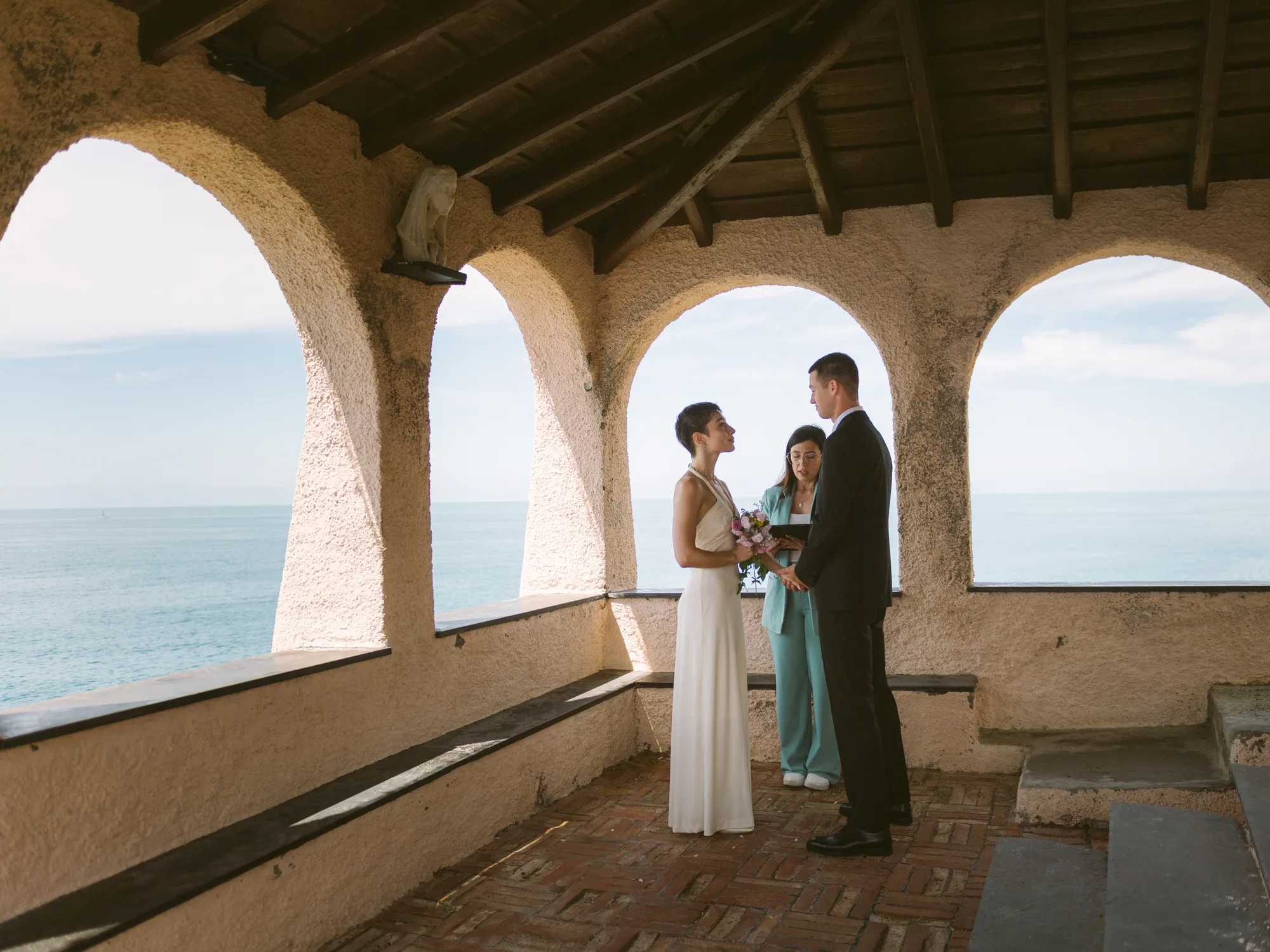 The bride in a white dress stands under an archway, holding a bouquet of flowers.