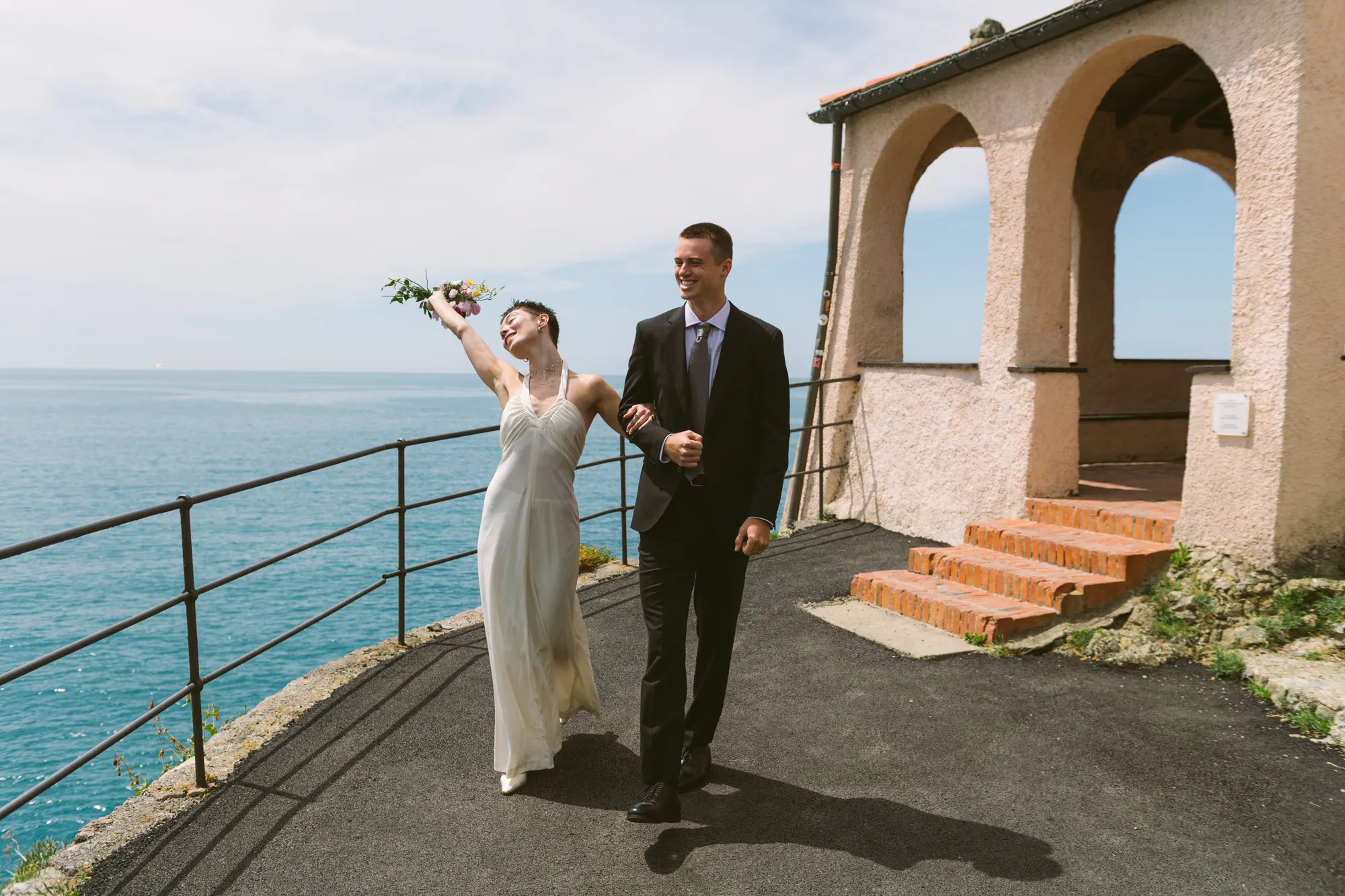 A bride in a white dress and a groom in a black suit are walking along a path near the sea.