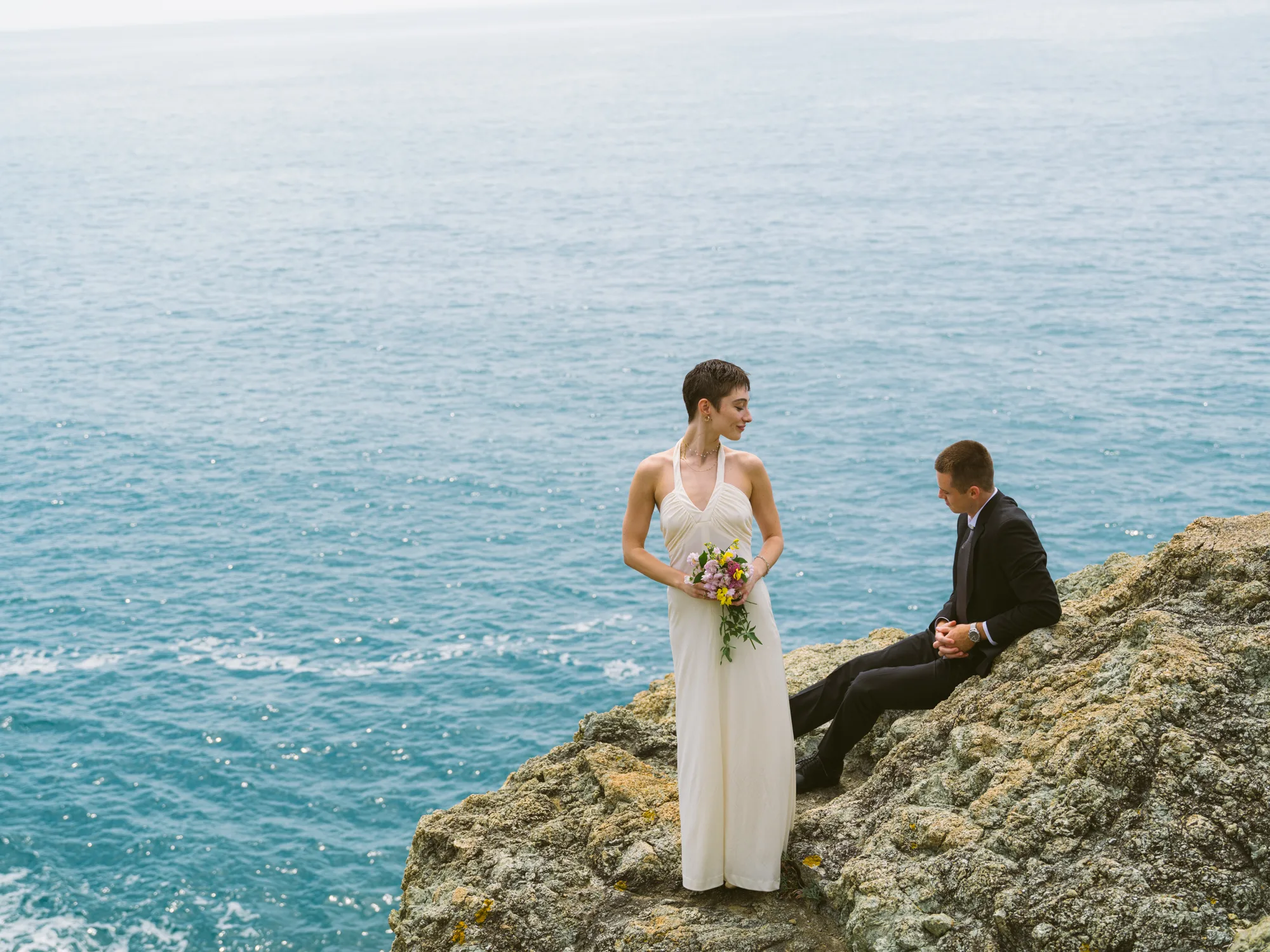 A bride in a white dress holds a bouquet of flowers, standing on a rocky cliff overlooking the ocean.