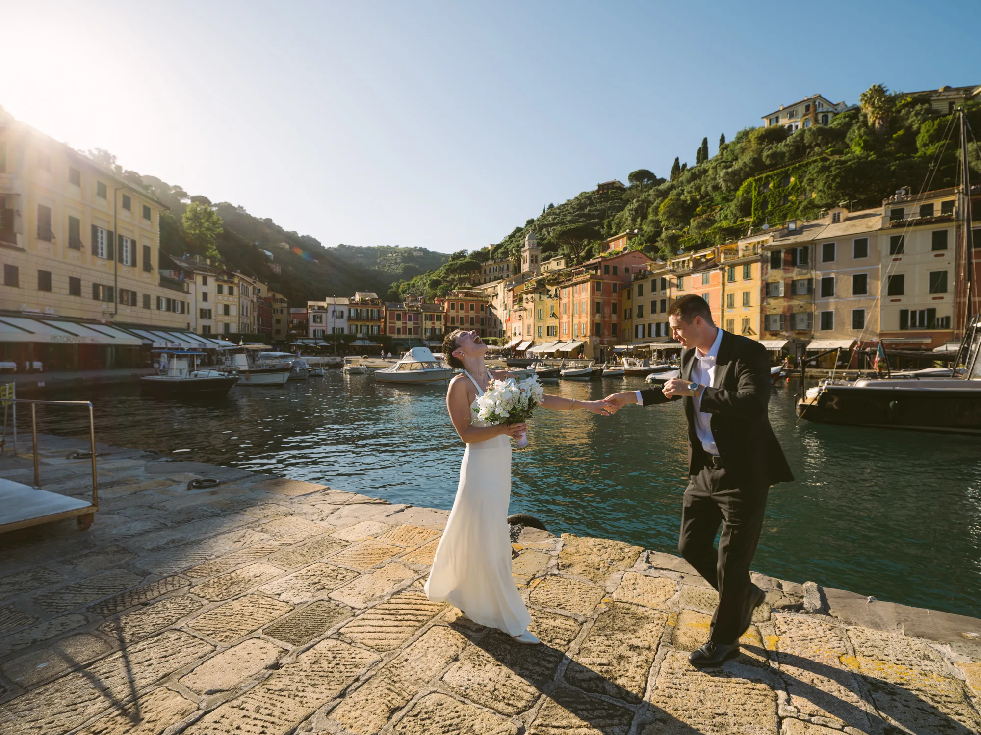 A bride in a white dress holds a bouquet of white flowers as she walks along a stone path near a Portofino harbor.