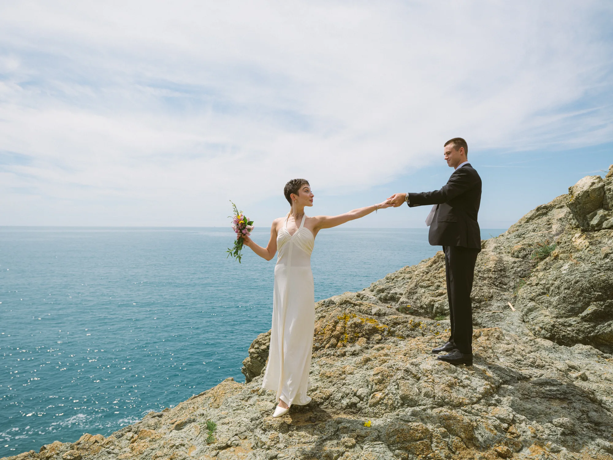 A bride in a white dress holds hands with a groom in a black suit on a rocky cliff overlooking the ocean.