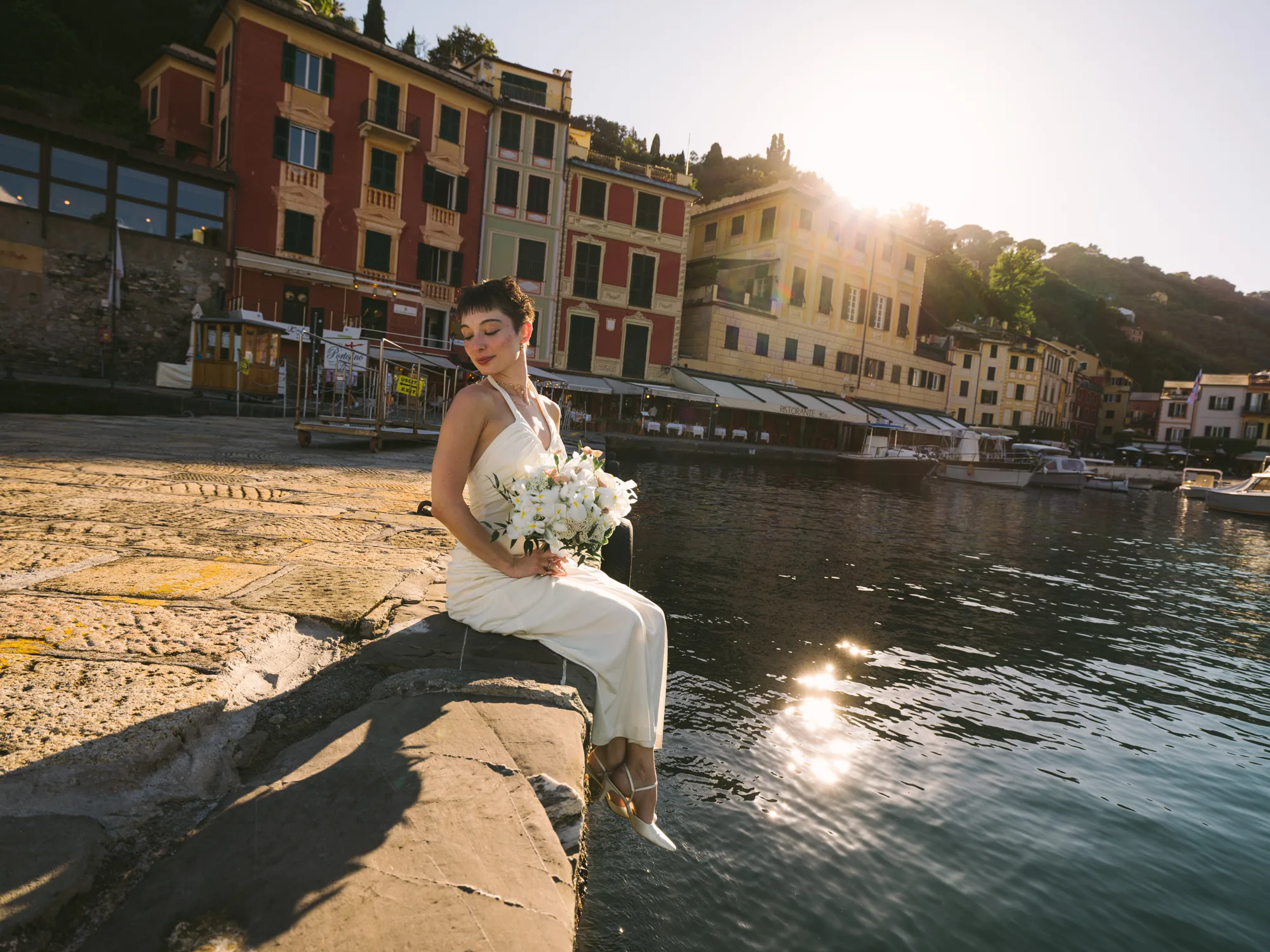 A bride in a white dress sits on a stone ledge by a body of water, holding a bouquet of white flowers in Portofino Italy