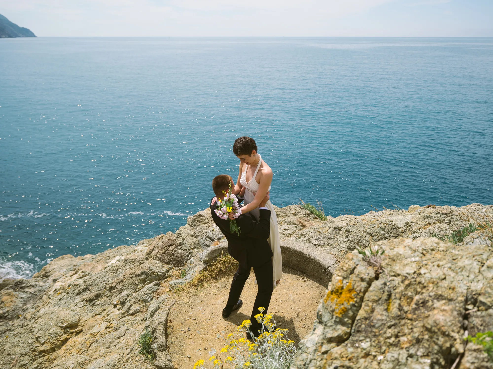 A bride in a white dress is standing on a rocky outcrop overlooking the Ligurian sea