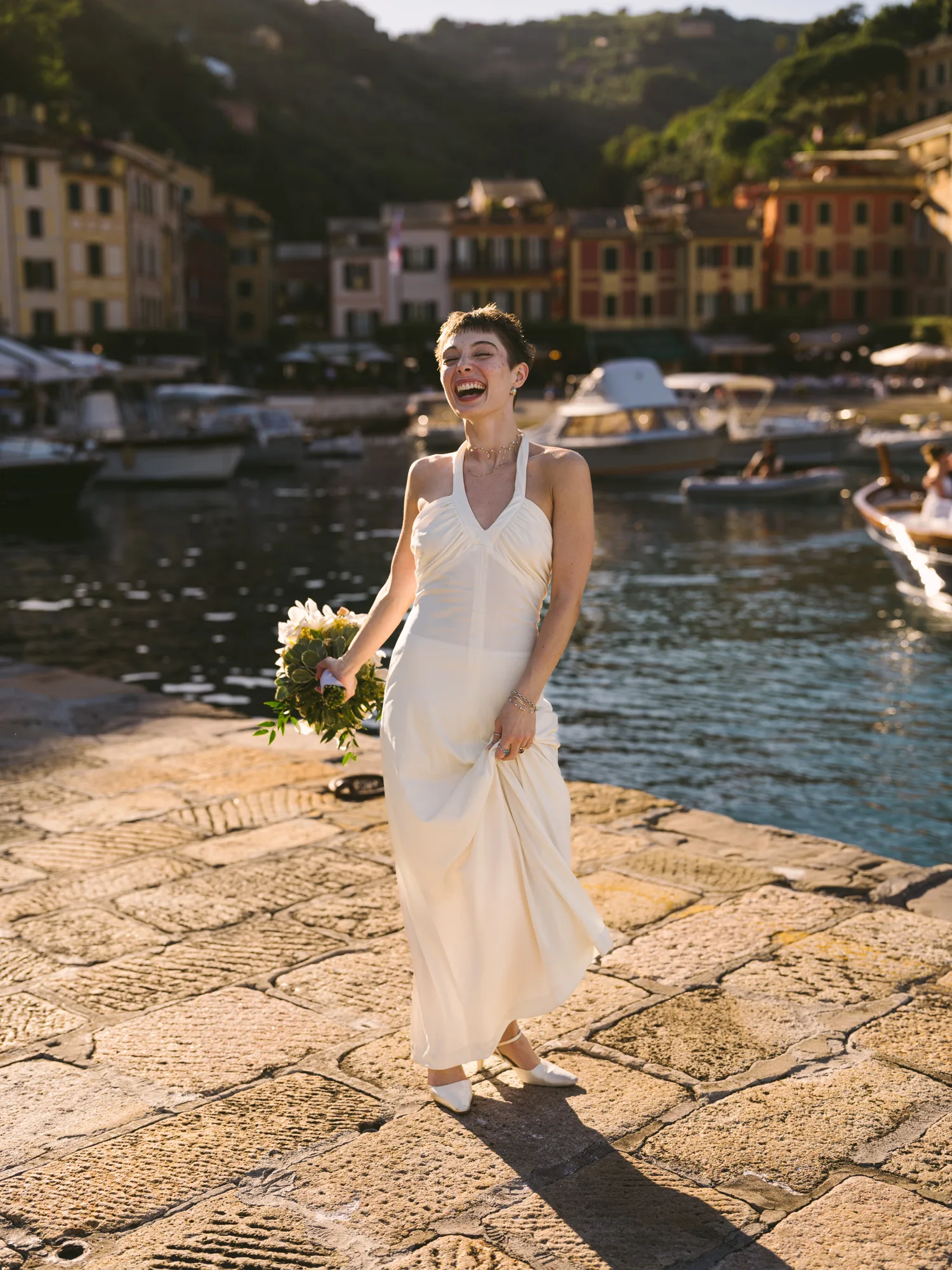 A bride laughing in a bridal dress stands on a cobblestone path by a body of water, holding a bouquet of flowers in Portofino Italy