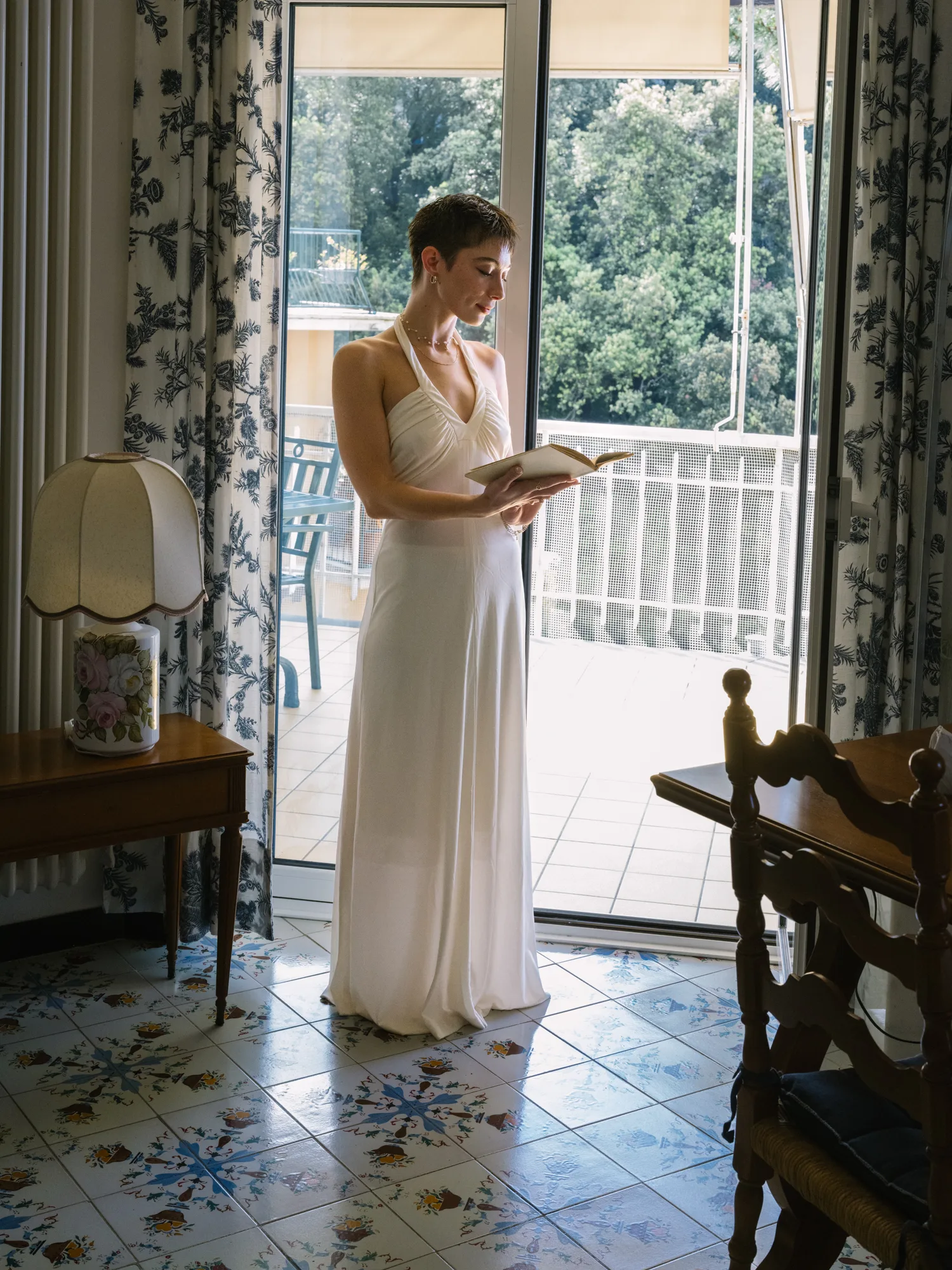 A bride in a white dress stands indoors, holding a book.