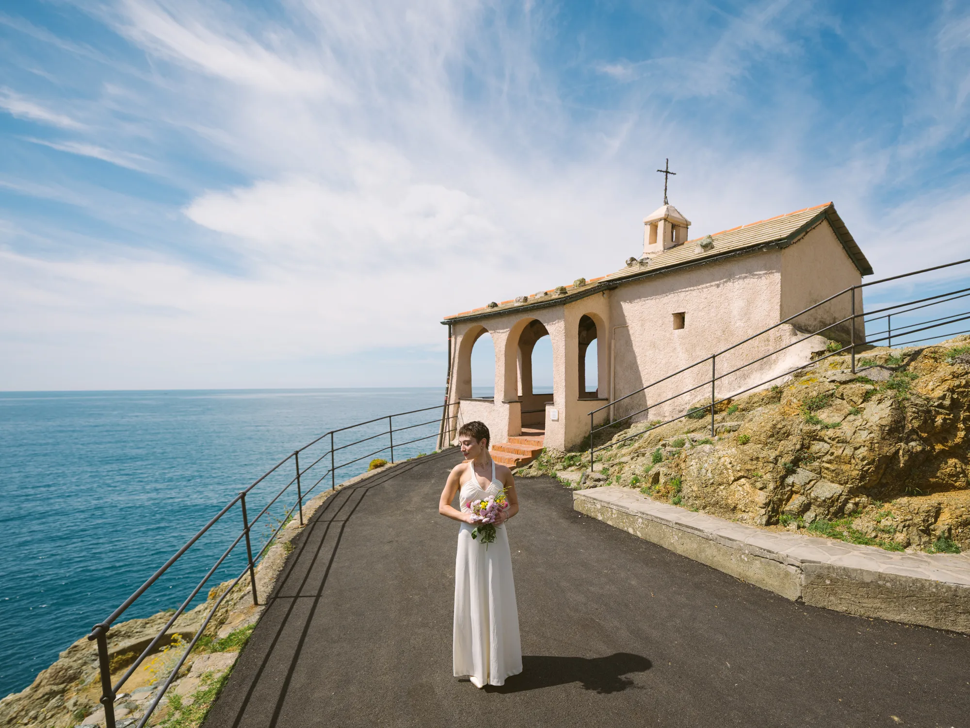 A bride in a white dress stands on a paved path near a small, arched building with a cross on top, overlooking the ocean.