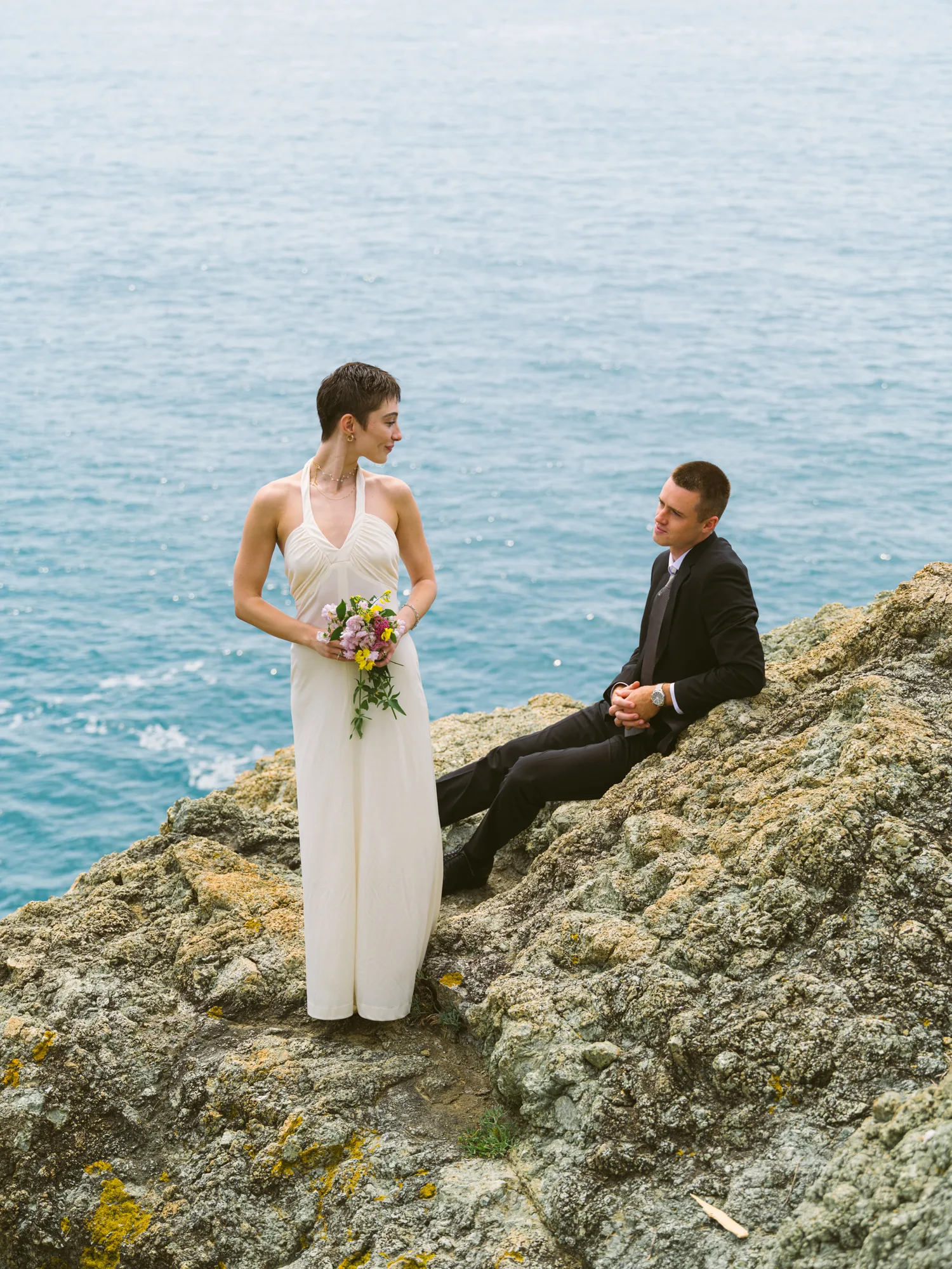 A bride in a white dress stands on rocky terrain, holding a bouquet of flowers.