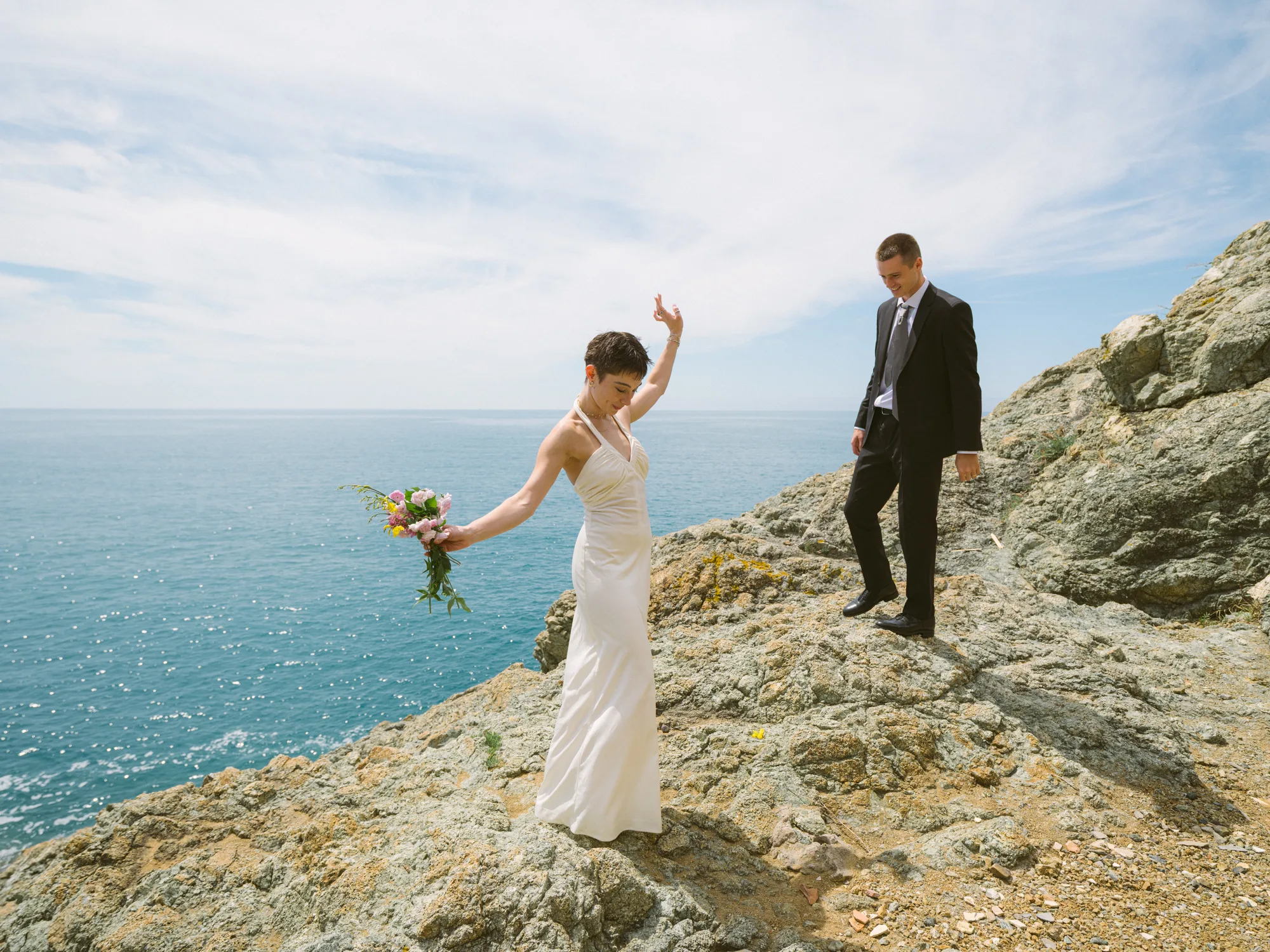 A bride in a white dress stands on rocky terrain, holding a bouquet of flowers with the groom