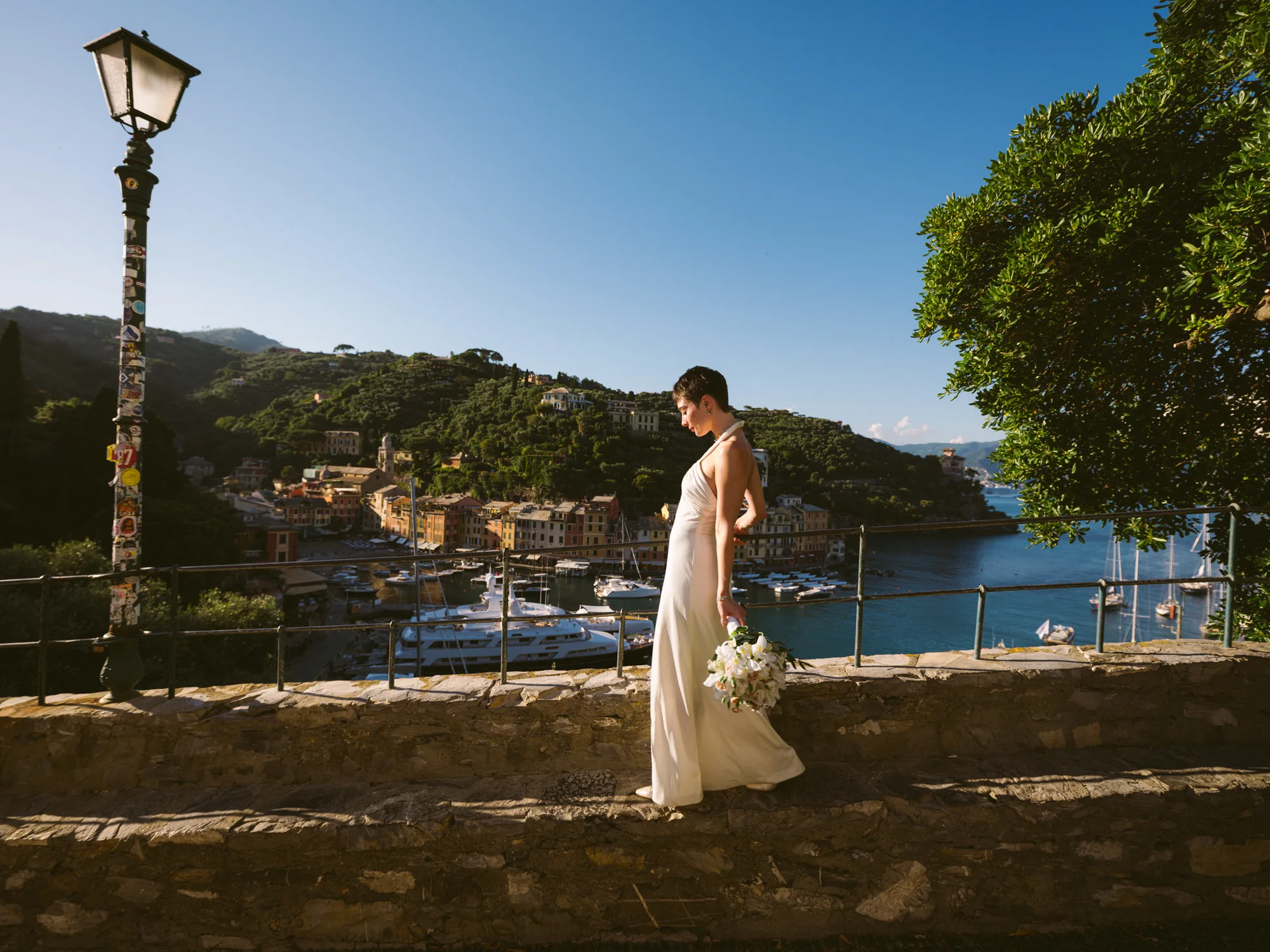 A bride in a white dress stands on a stone wall, holding a bouquet of white flowers In Portofino