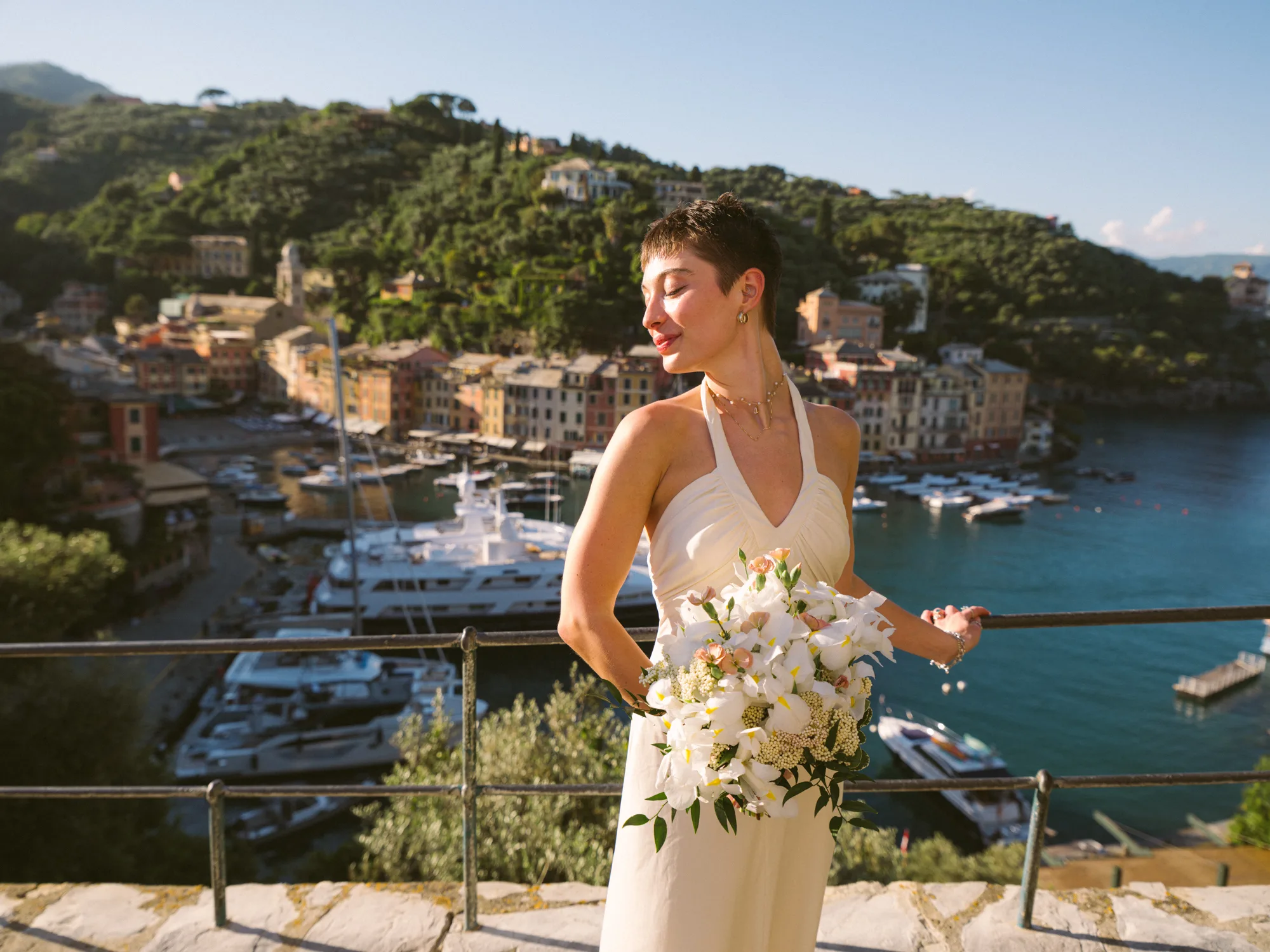 A bride in a white dress stands on a stone wall, holding a bouquet of white flowers with green leaves in Portofino