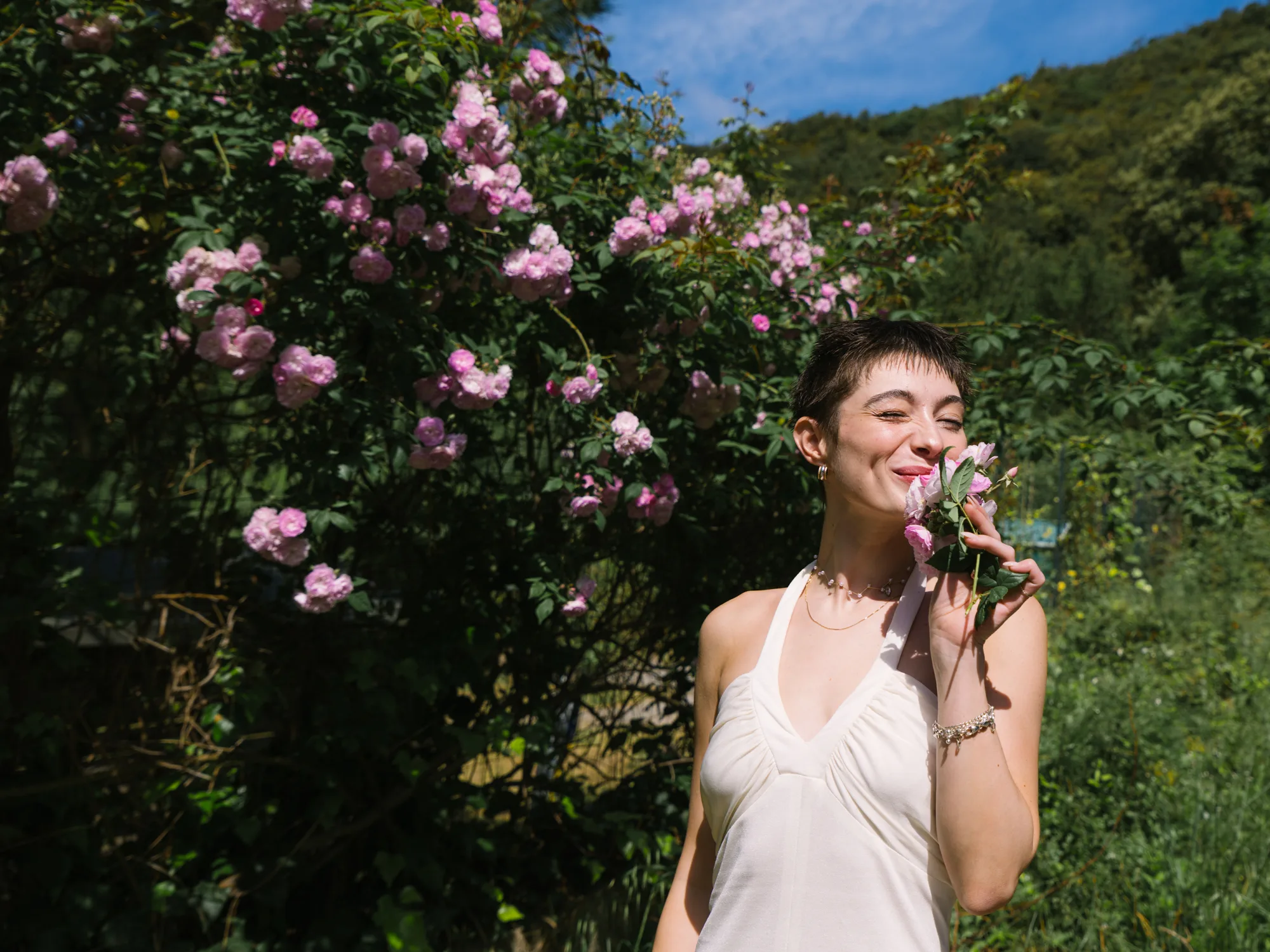 A bridein a white sleeveless top stands in front of a bush of pink roses.