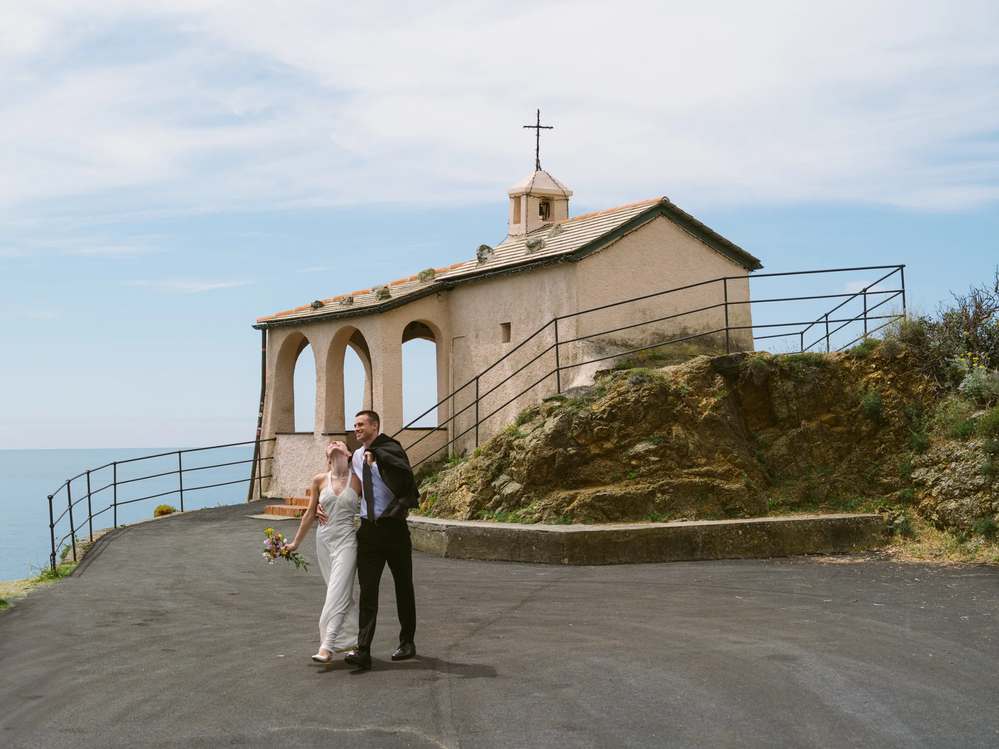 A couple is dancing in front of a small, rustic chapel with a cross on top, set against a backdrop of a rocky cliff and a clear sky.