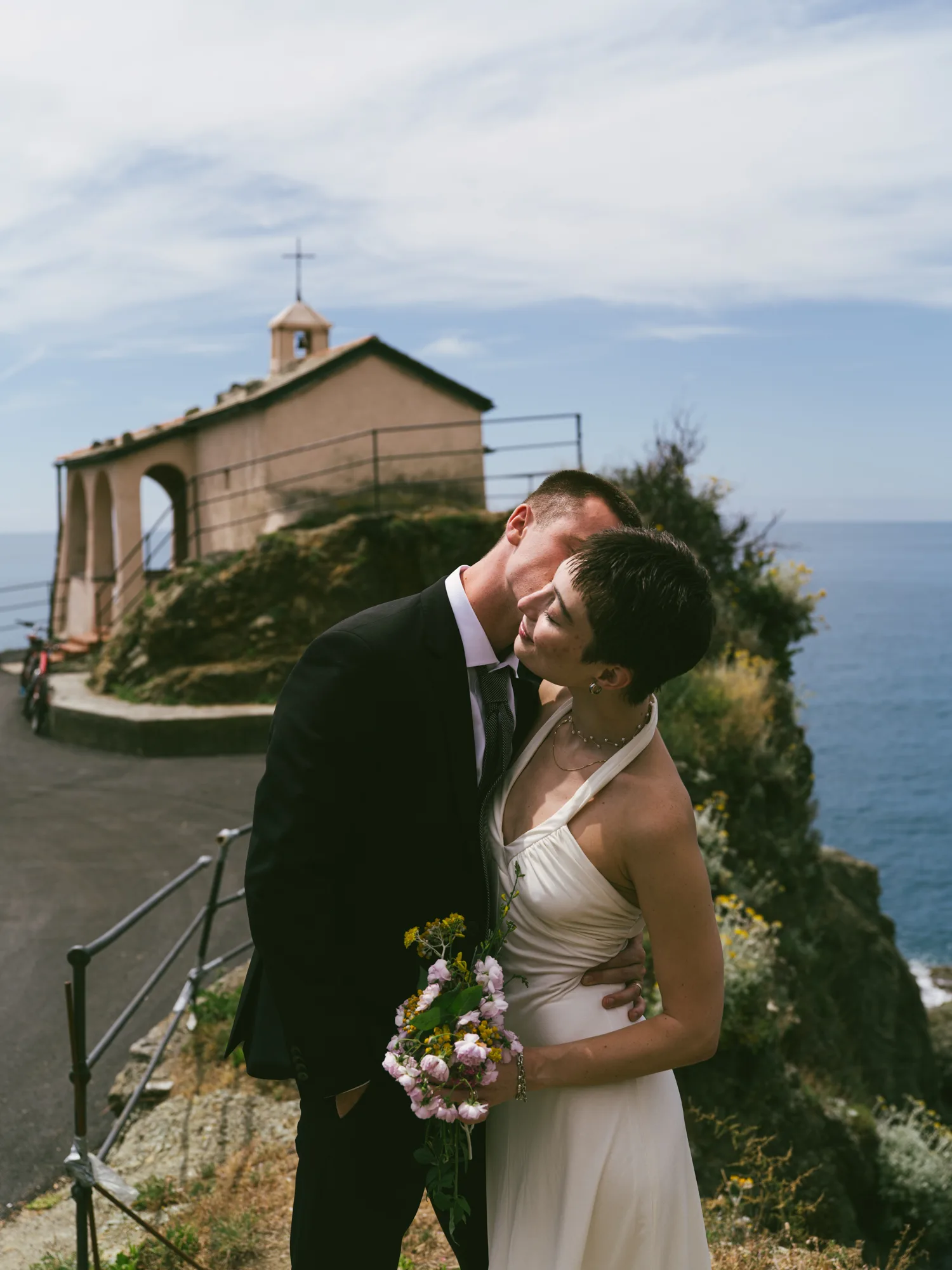 A couple is embracing on a scenic coastal path, with a small chapel and a cross visible in the background.