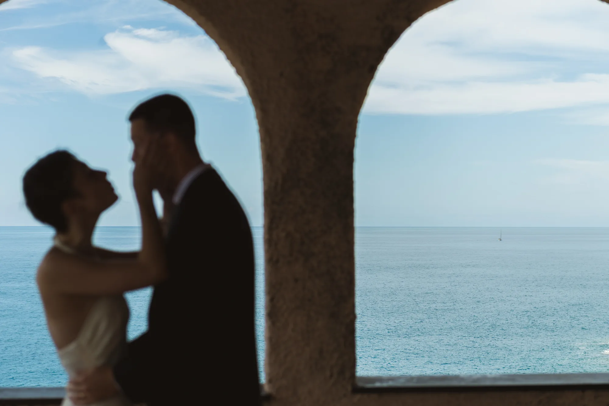 A couple is embracing under an archway, with the ocean and sky visible in the background.