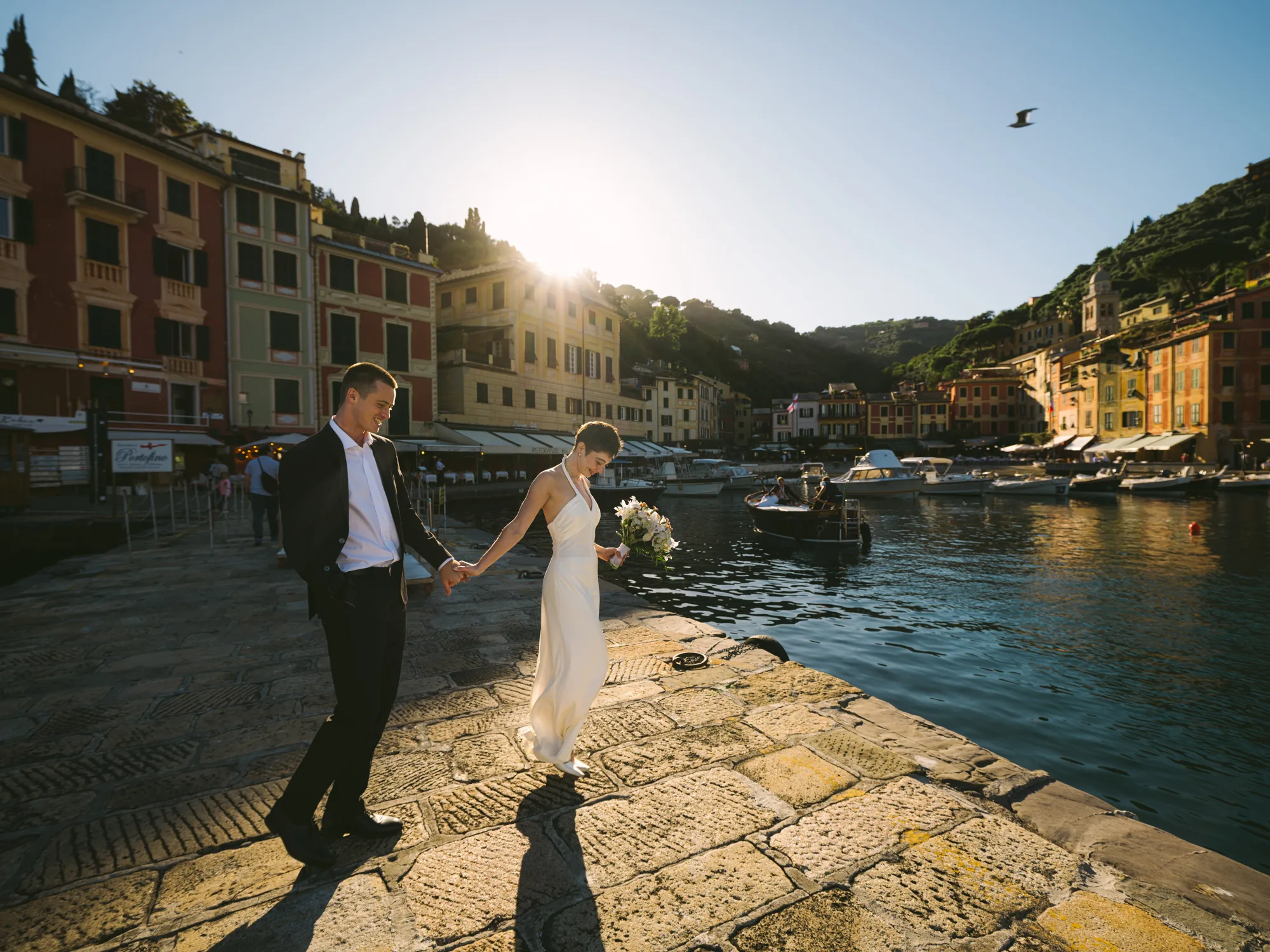 A couple is holding hands on a stone-paved walkway by a body of water in Portofino