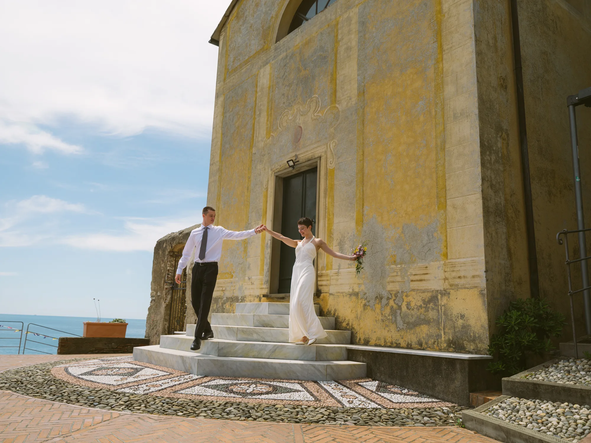 A couple is holding hands and walking down a set of stairs in front of Ex Oratorio di Sant’Erasmo church in Bonassola