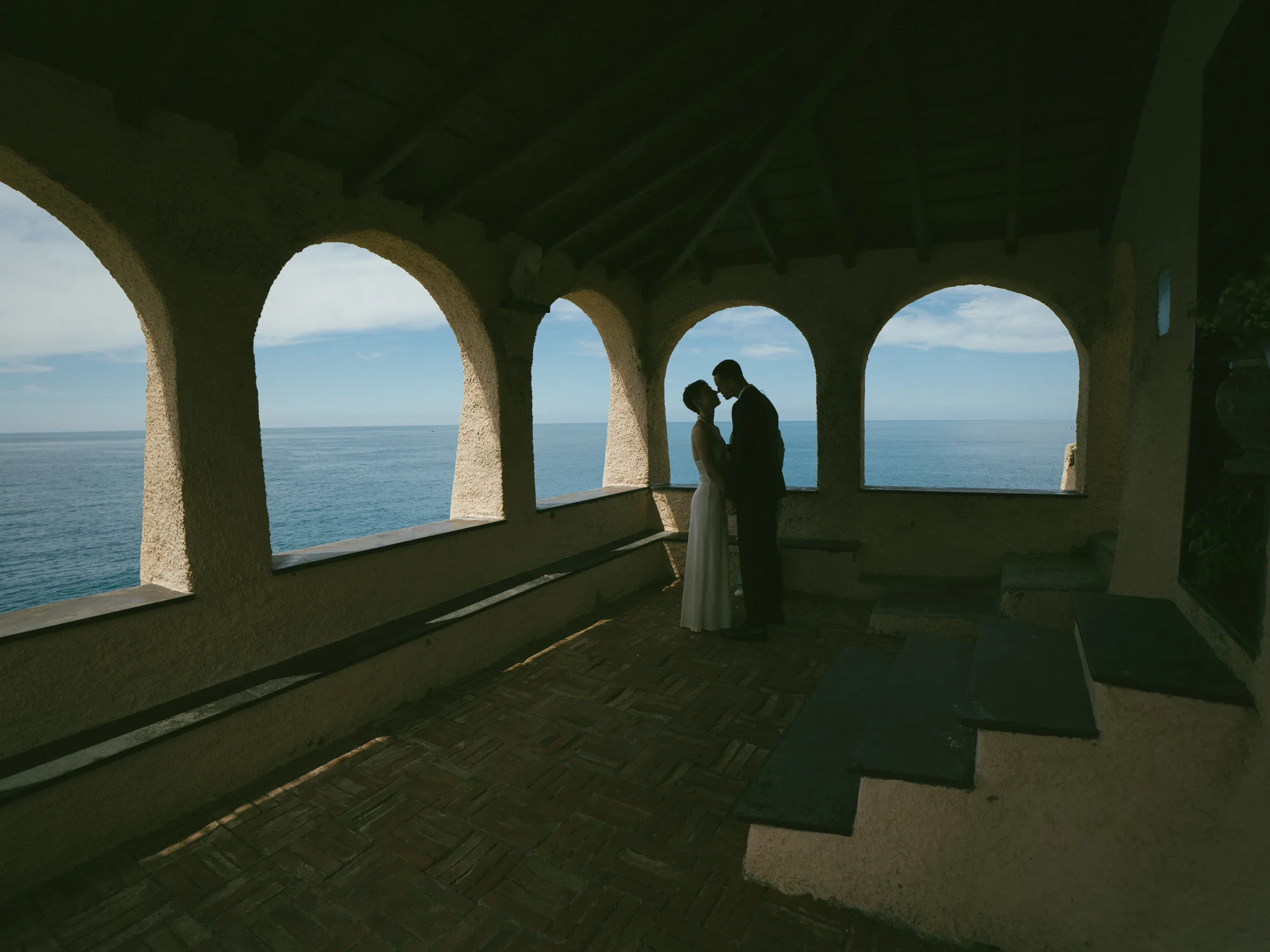 A couple is kissing in a covered outdoor area with arches and a view of the ocean.