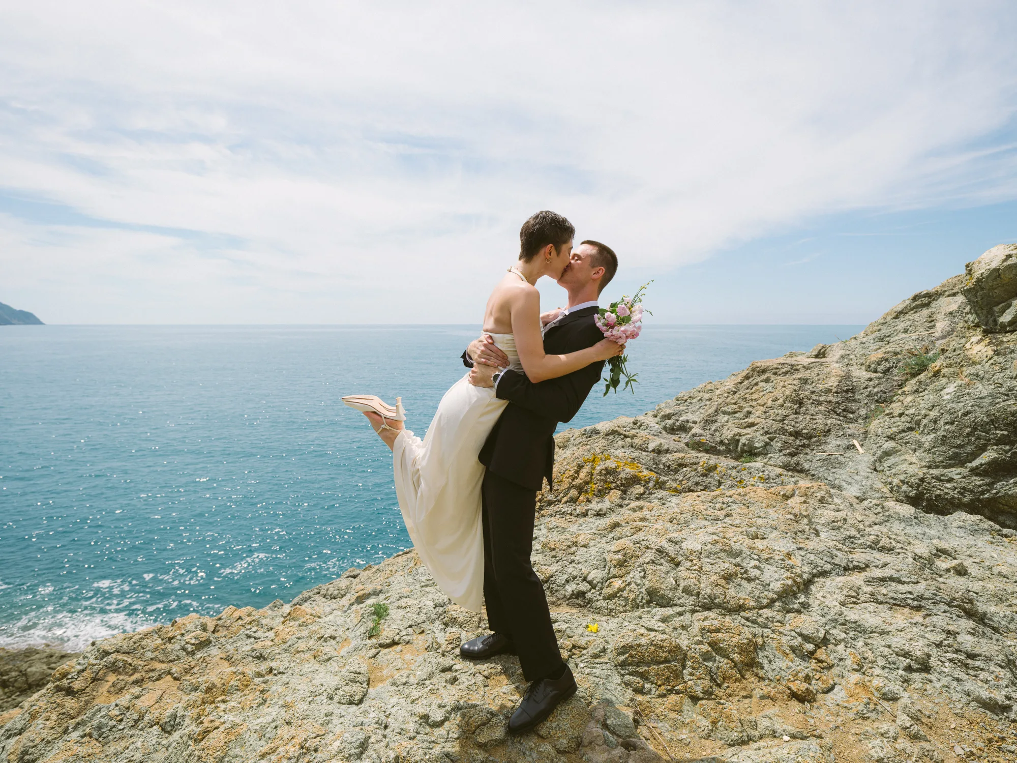 A couple is kissing on a rocky cliff overlooking the ocean.