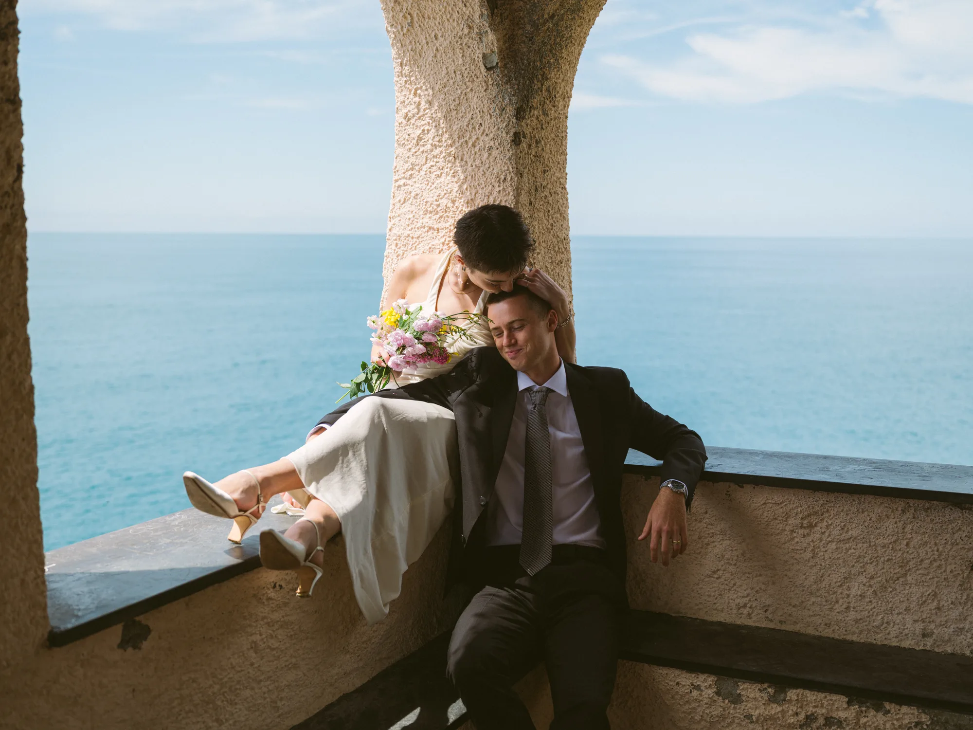 A couple is sitting on a ledge overlooking the ocean.