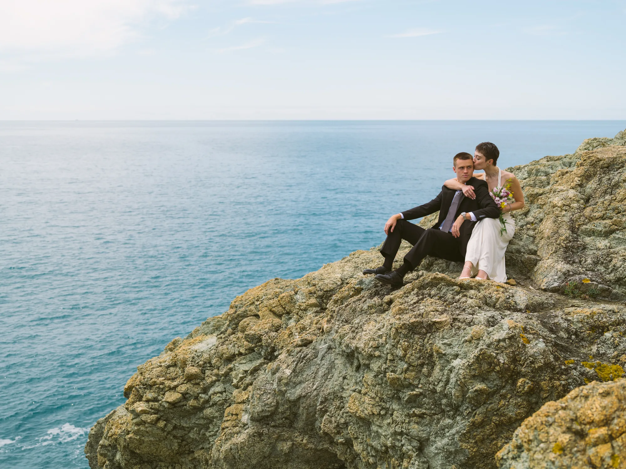 A couple is sitting on a rocky cliff overlooking the ocean.