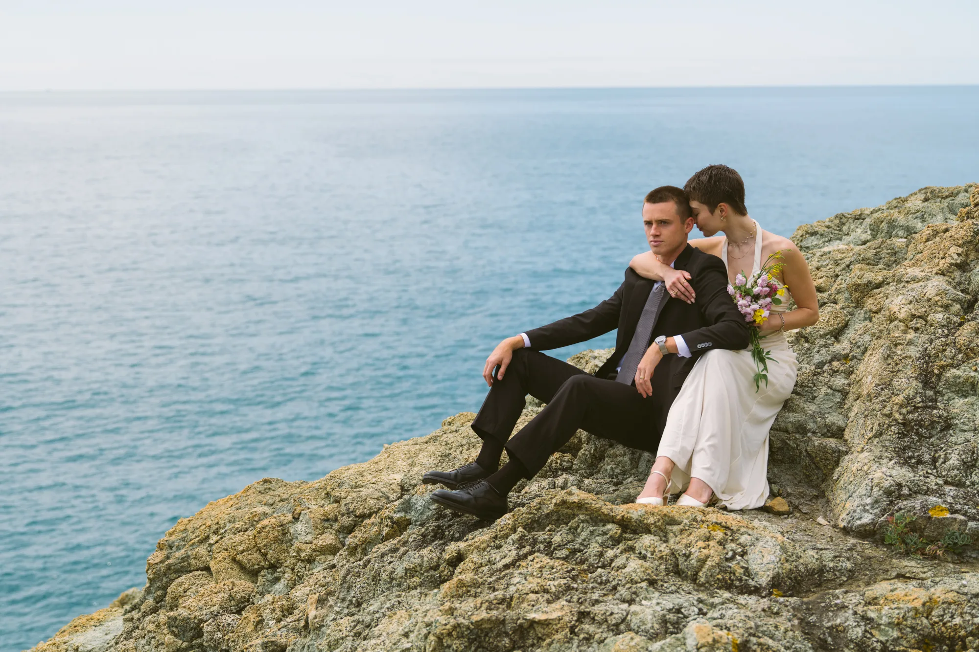 A couple is sitting on a rocky cliff overlooking the ocean.