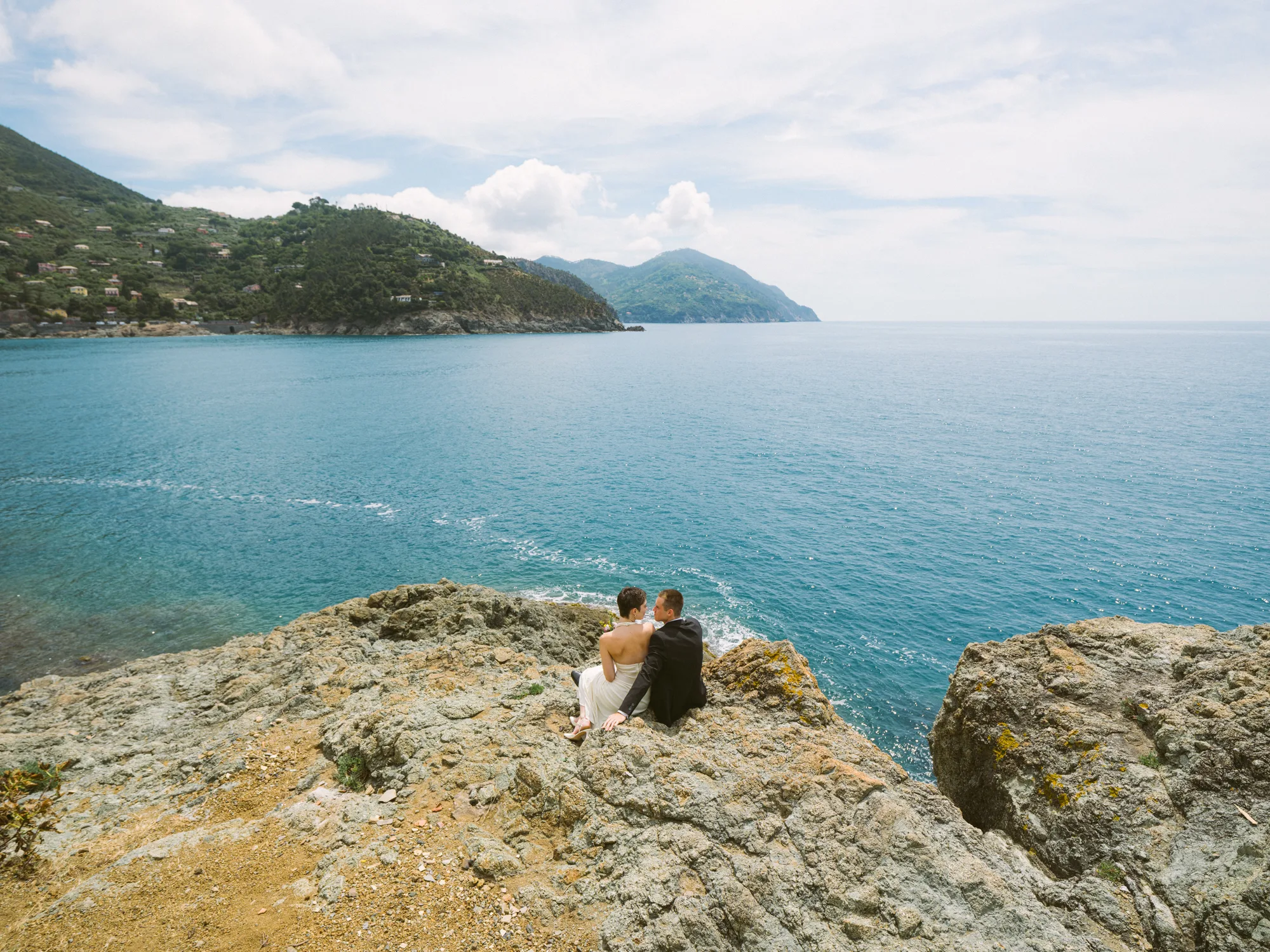 A couple is sitting on a rocky cliff overlooking a vast, turquoise sea.