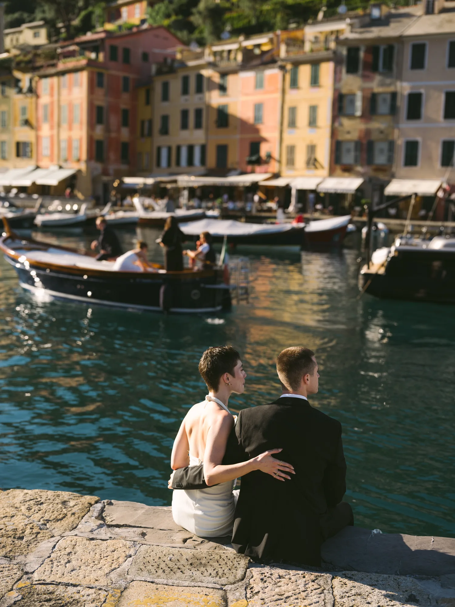 A couple is sitting on a stone wall by the water, with a boat and colorful buildings in the background in Portofino
