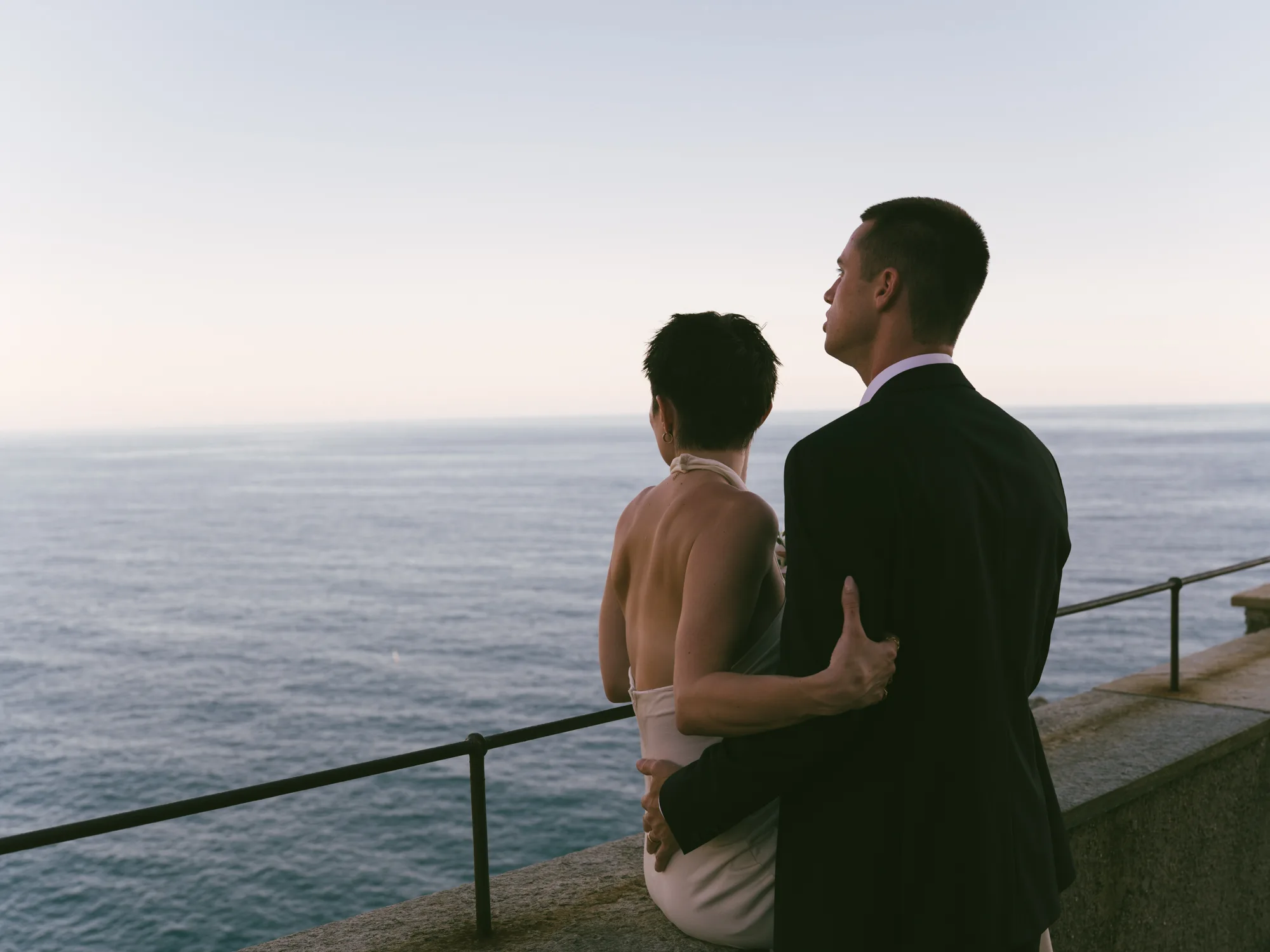A couple is standing on a balcony overlooking the ocean, with the man in a dark suit and the woman in a white dress overlooking Portofino beach