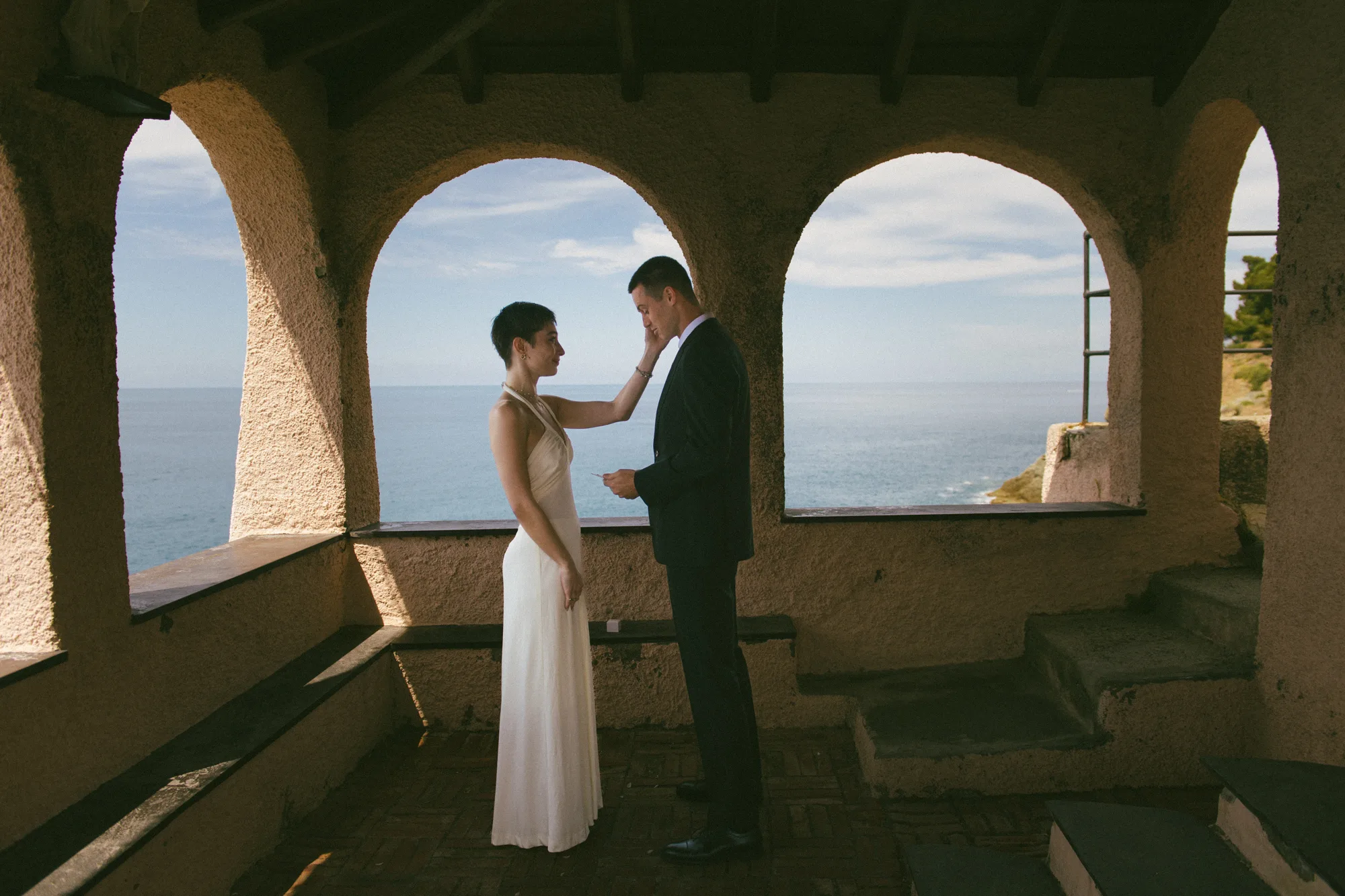 A couple stands on a balcony overlooking the ocean, with the woman in a white dress and the man in a dark suit.