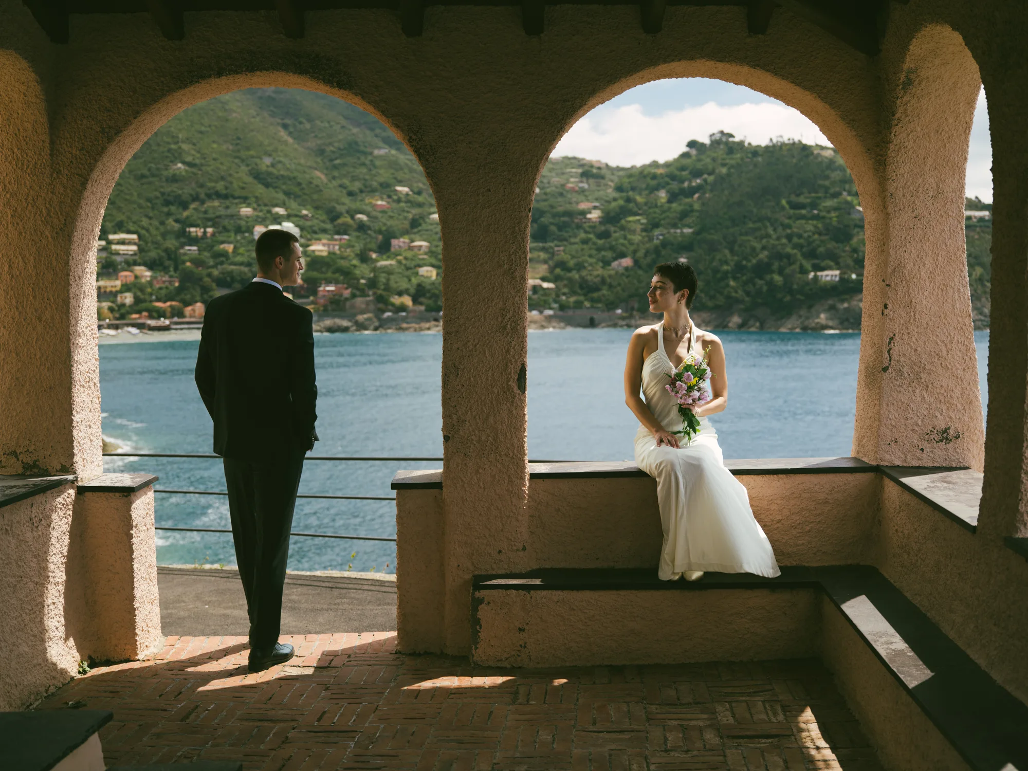 A couple stands on a balcony overlooking a scenic coastal view, with the man in a dark suit and the woman in a white dress holding a bouquet of flowers.