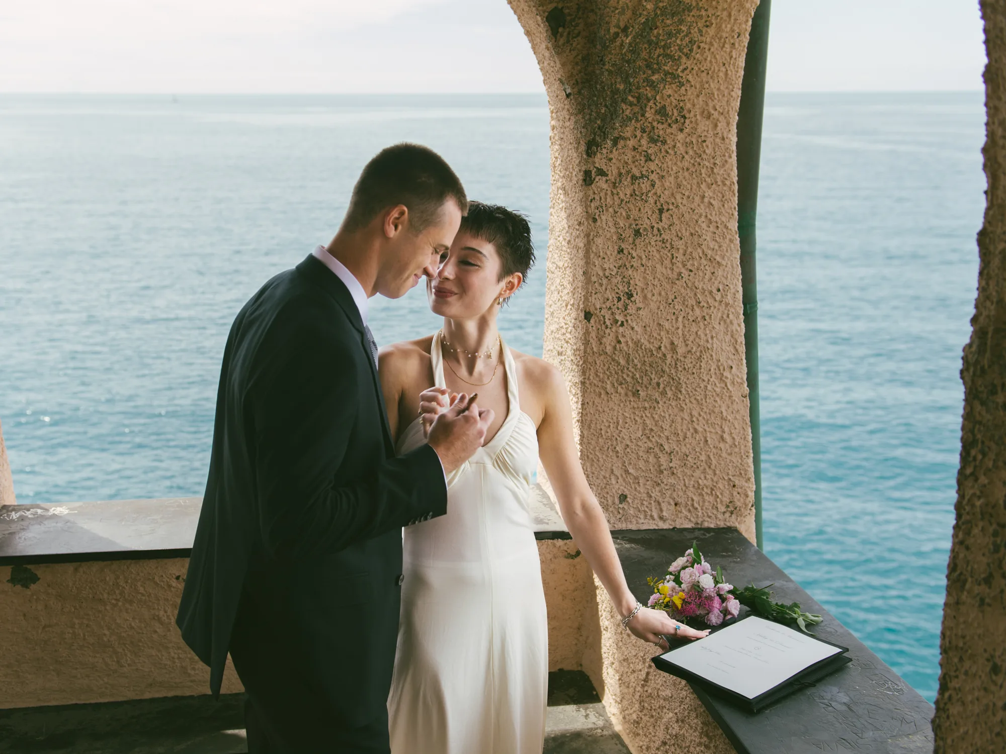 A couple stands close together, with the man in a dark suit and the woman in a white dress.