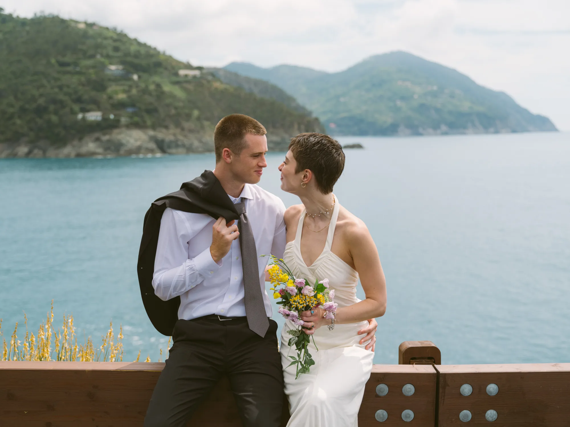 A couple stands close together, the man in a white shirt and black pants, the woman in a white dress holding a bouquet of flowers.
