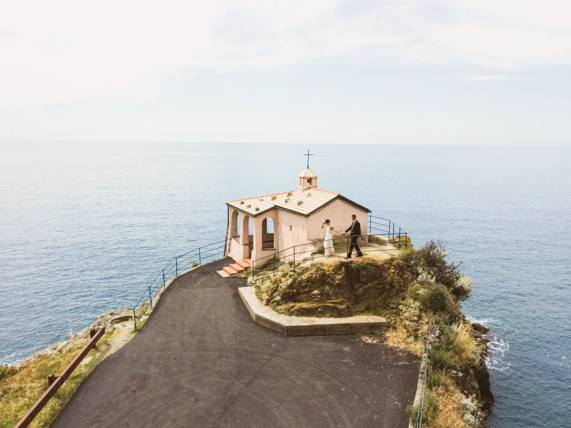 A couple stands on a narrow path leading to a Cappella della Madonnina della Punta perched on a rocky cliff overlooking the ocean.