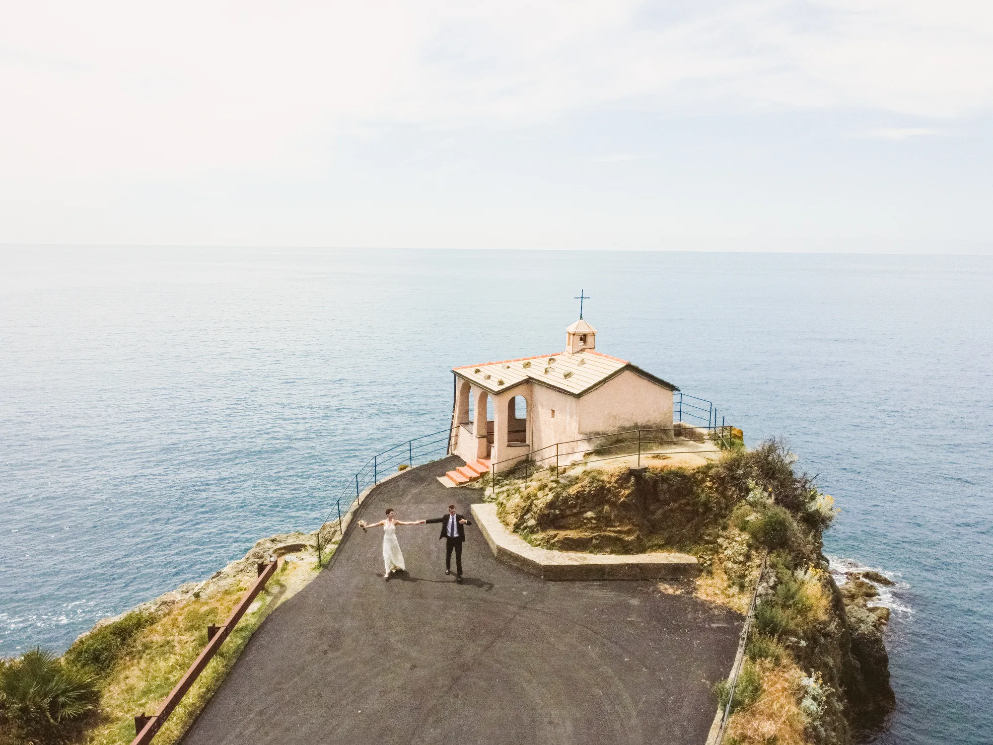 A couple stands on a paved path overlooking the ocean, with a Cappella della Madonnina della Punta chapel-like structure with a cross on top visible in the background.