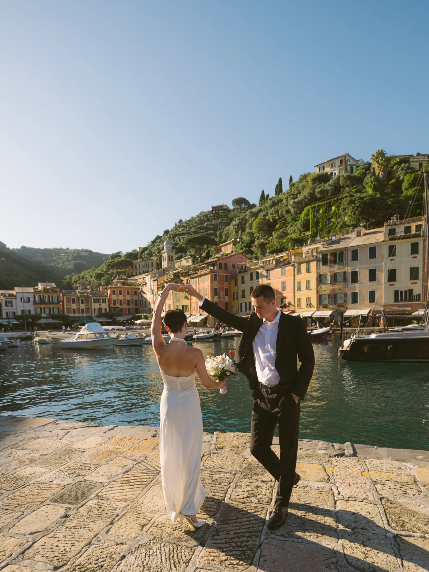 A couple stands on a stone-paved dock by a picturesque harbor, with colorful buildings and lush greenery in the background in Portofino