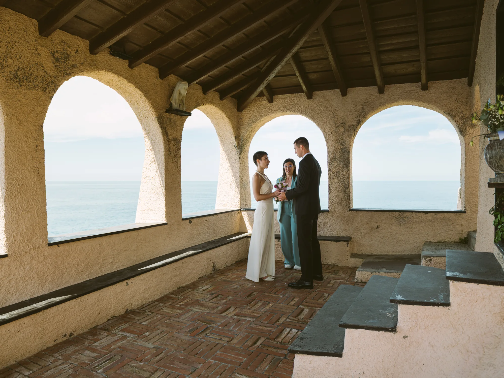 A couple stands under an arched, rustic structure with a view of the ocean.