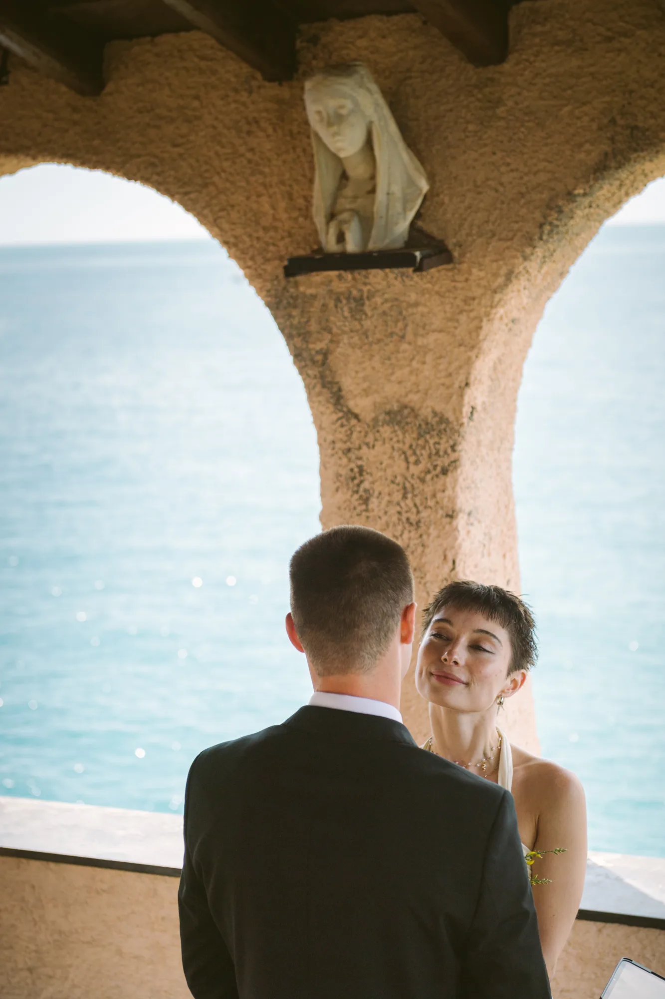 A couple stands under an archway with a statue above them, facing each other.