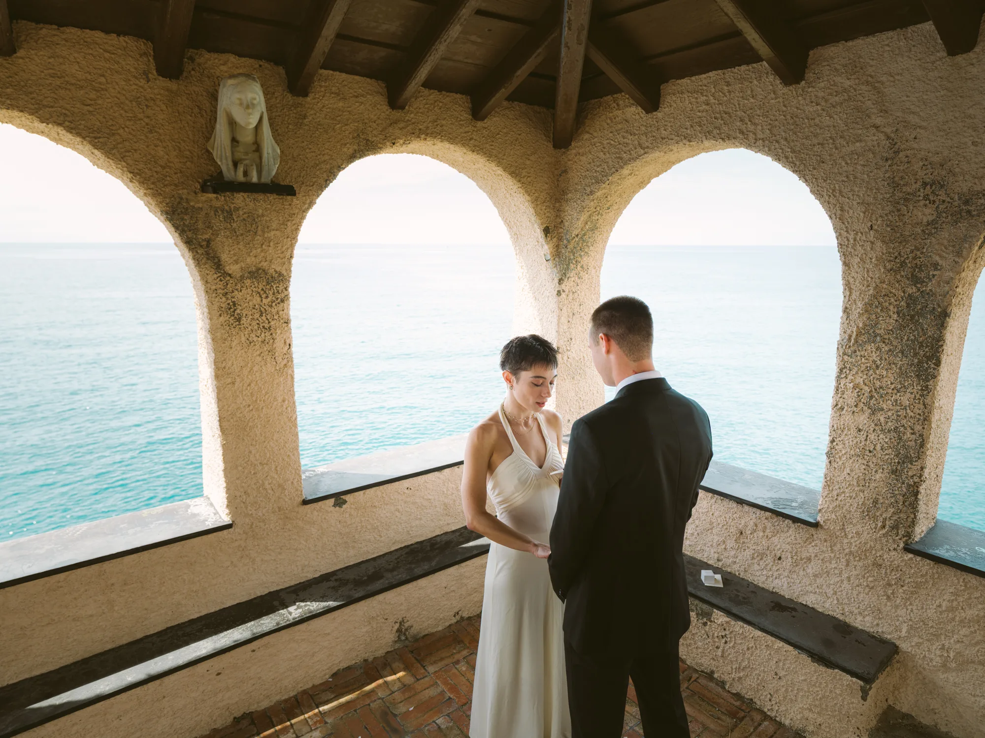 A couple stands under an archway with a view of the ocean.