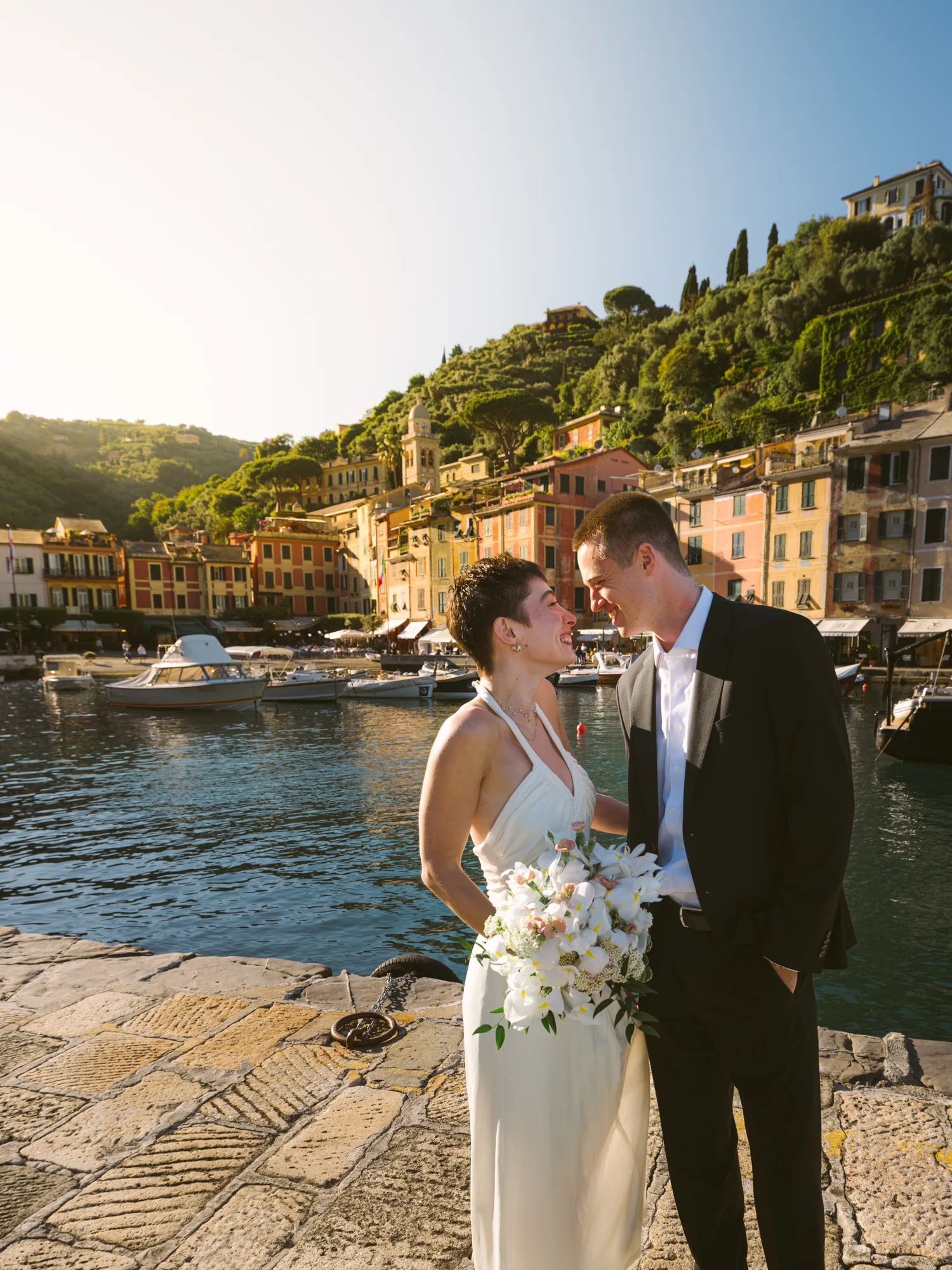 The photograph features a bride and groom standing on a stone-paved dock by a body of water in Portofino