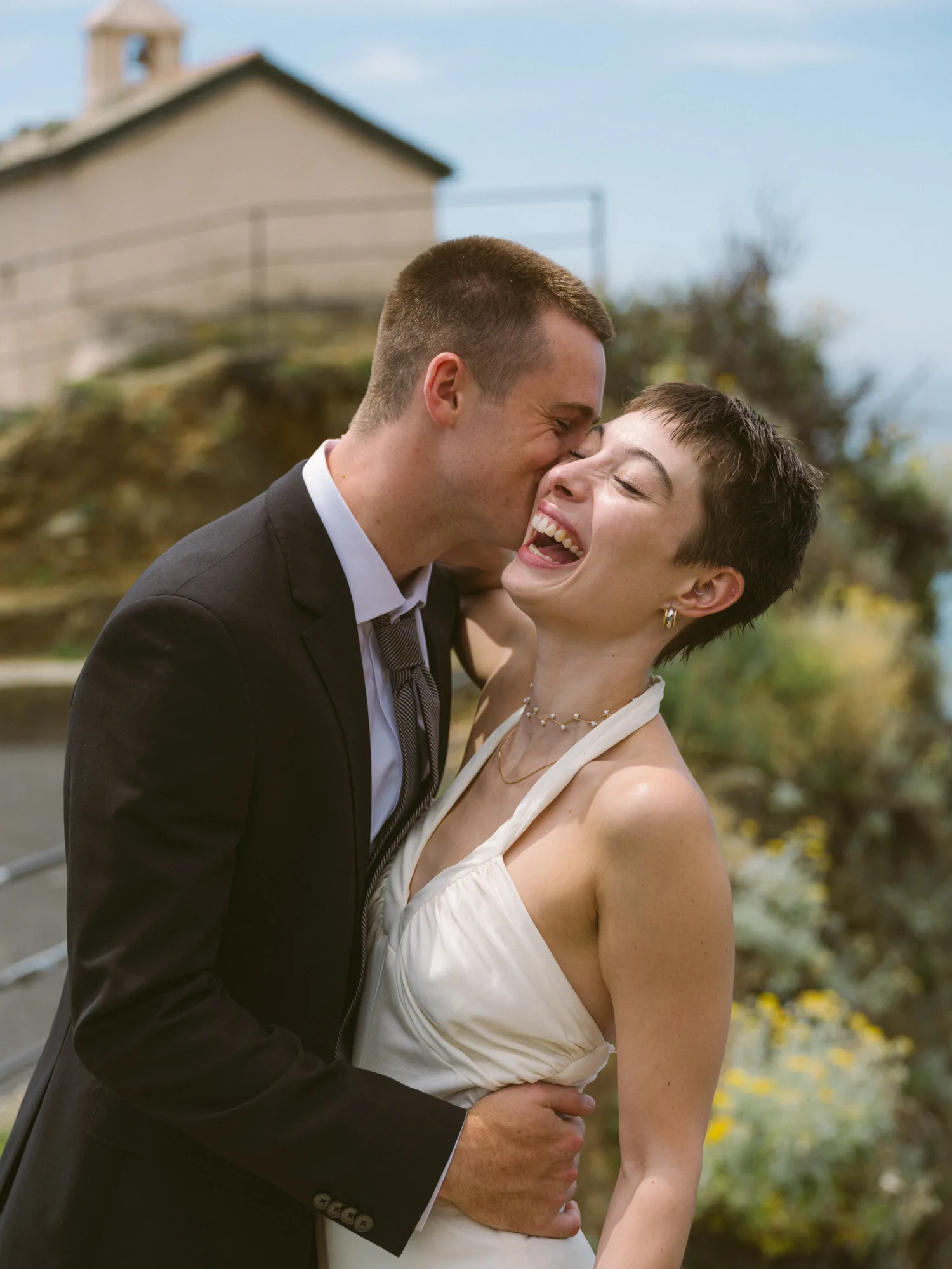 A groom in a black suit and a bride in a white dress are embracing outdoors.