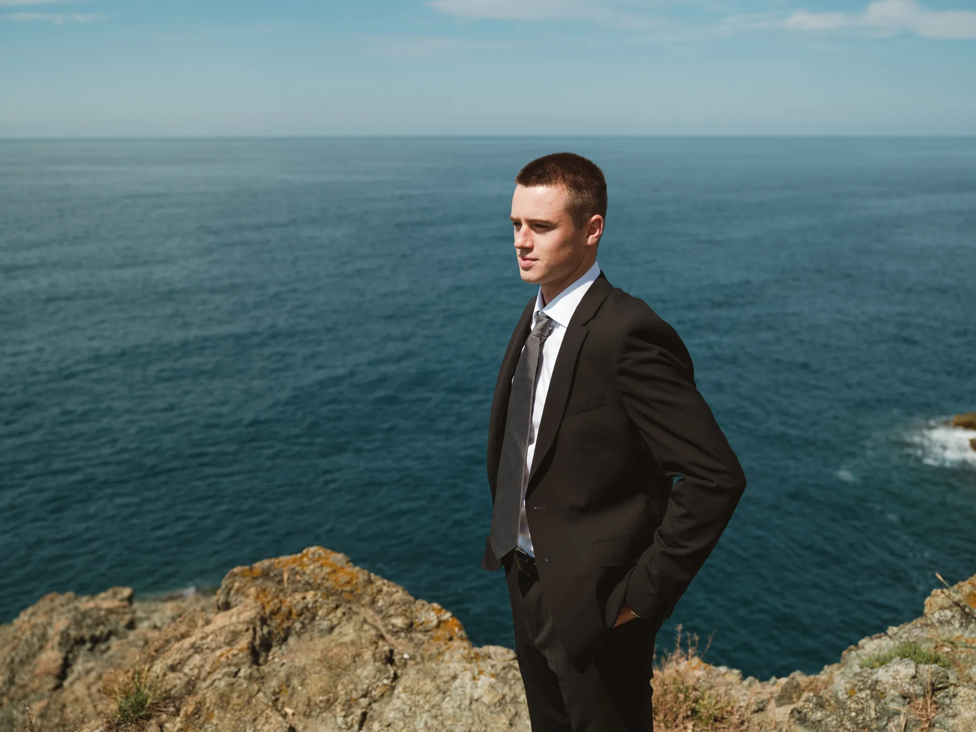 A groom in a black suit stands on a rocky cliff overlooking a vast, calm ocean.