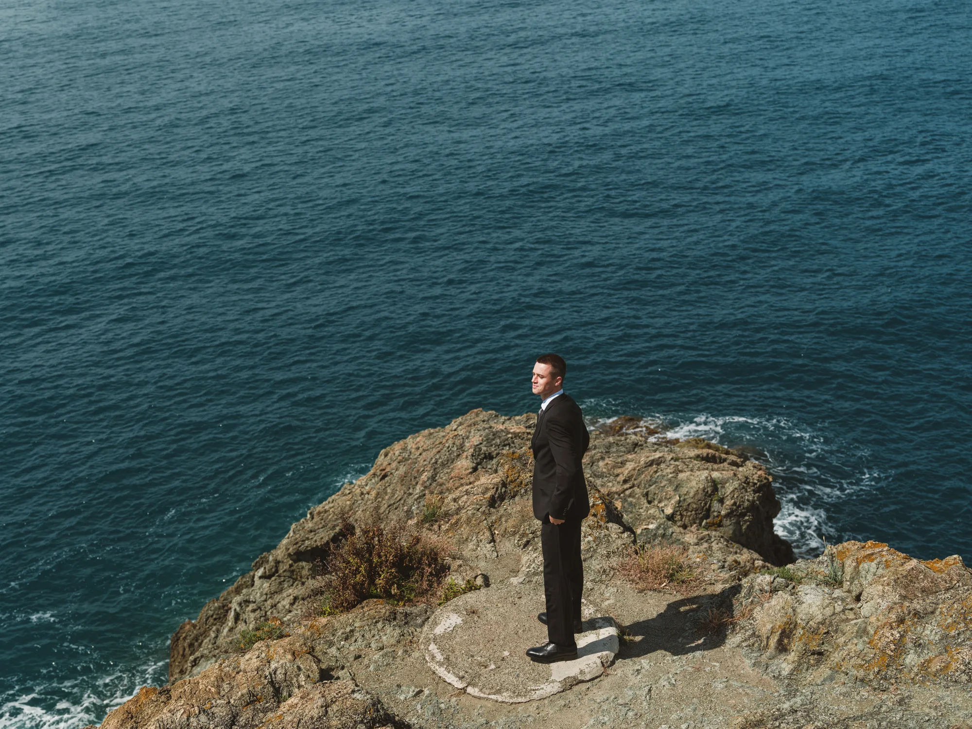 A groom in a black suit stands on a rocky outcrop overlooking the ocean.